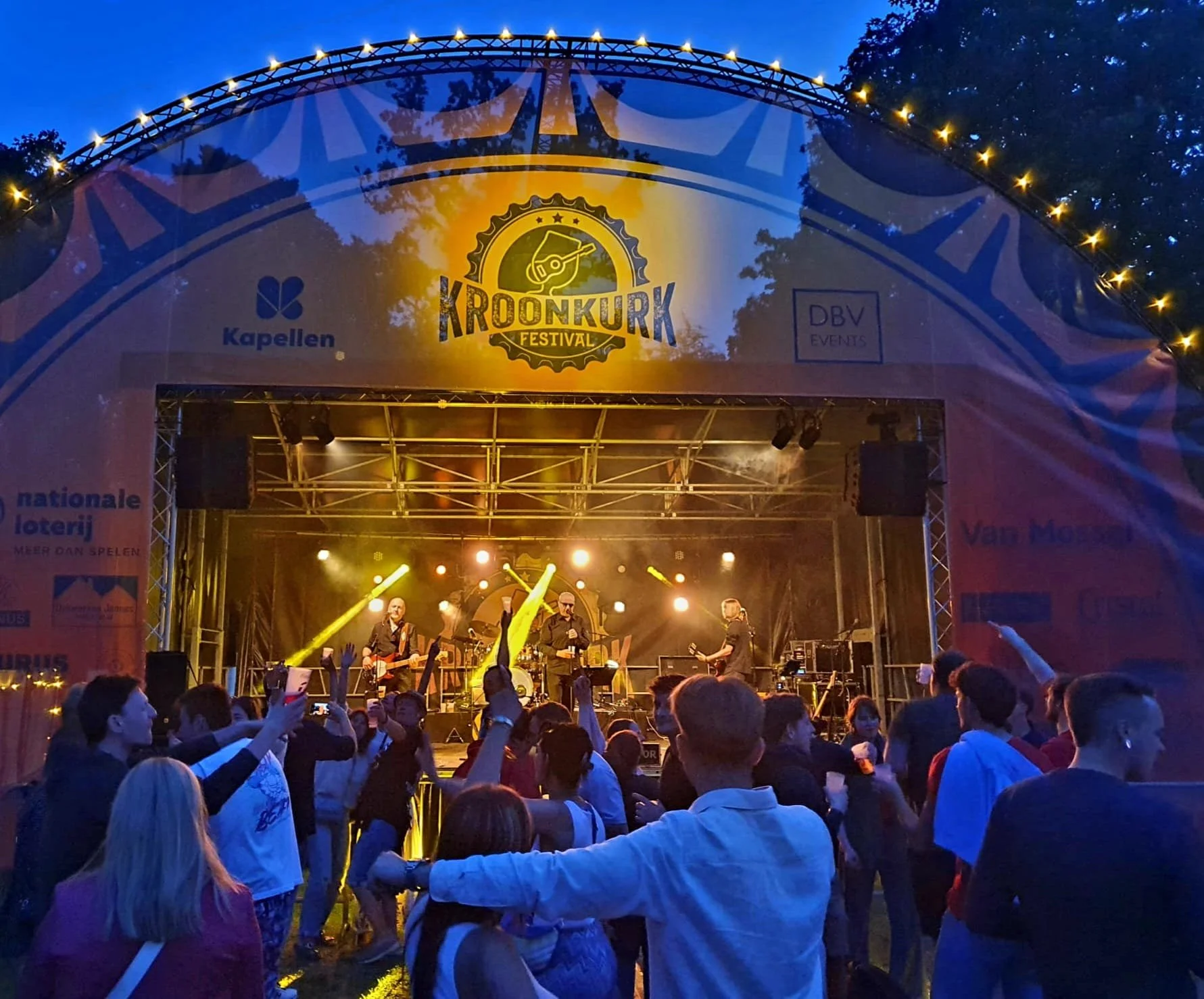 Een live muziekoptreden op het Kroonkurk Festival met een band op het podium en een publiek dat danst en fotografeert, onder een verlichte tent met het festivallogo bovenaan.