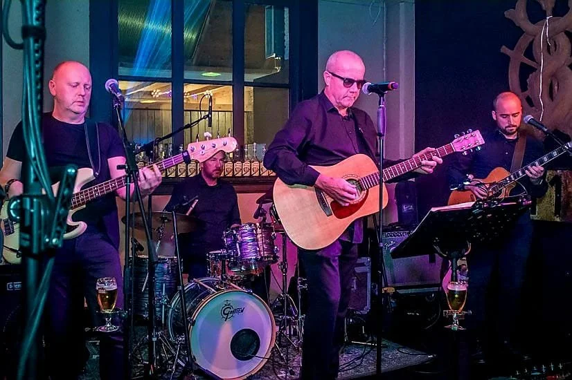 Vier muzikanten spelen live muziek in een bar of club, met gitaren en drums, onder kleurrijk podiumlicht.