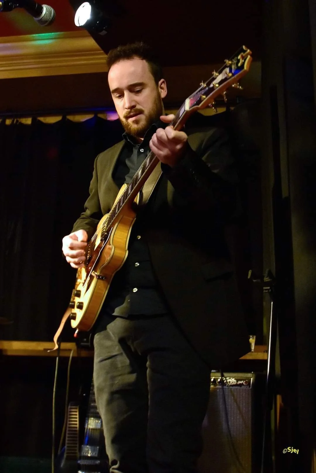 Man speelt elektrische gitaar op een podium in een donkere ruimte, met een versterker op de achtergrond.