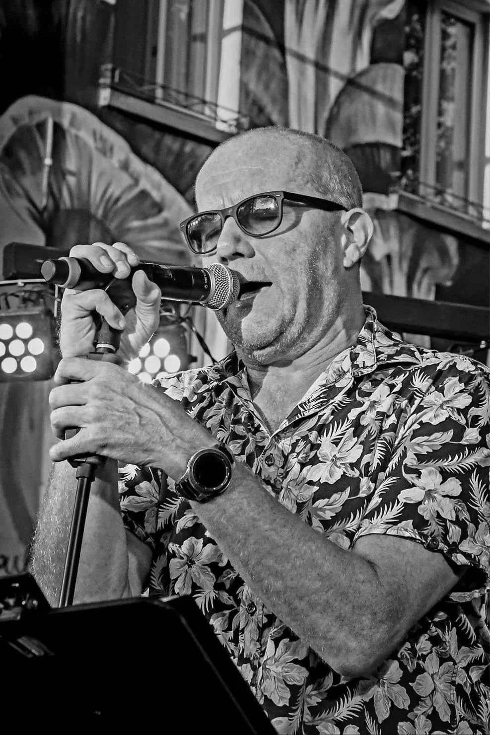 Zwarte en witte foto van een man met een kale hoofdhuid, donkere bril en een shirt met bloemmotief, die zingt of praat in een microfoon op een podium.
