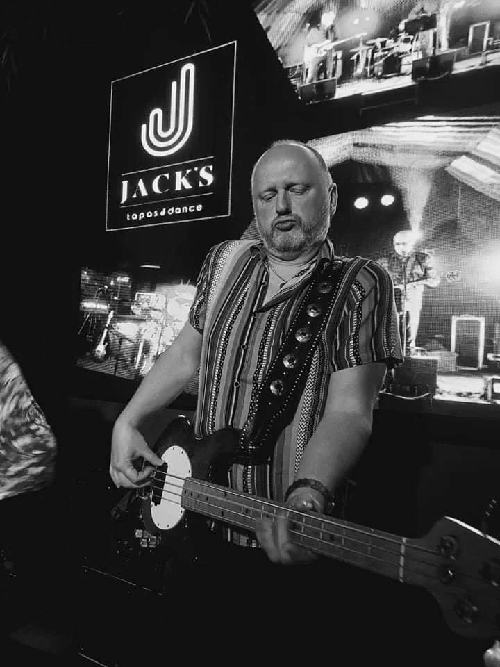 Een man die op een podium een elektrische basgitaar bespeelt, met een scherm op de achtergrond dat 'JACKS tapas & dance' toont.