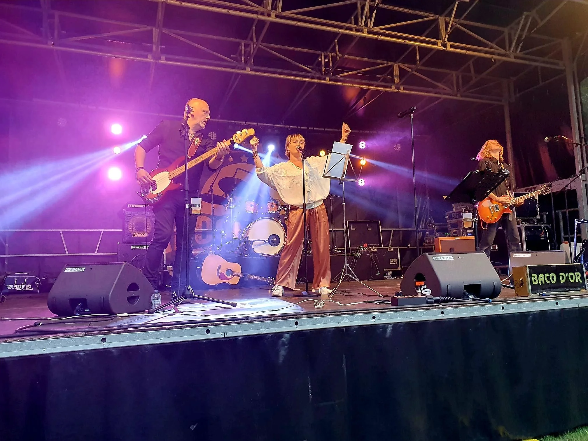 Een band speelt live op een podium met kleurrijke lichtstralen en een drummer op de achtergrond. Er zijn drie muzikanten: een man met een basgitaar, een vrouw die zingt en gitaar speelt, en een andere vrouw met een elektrische gitaar.