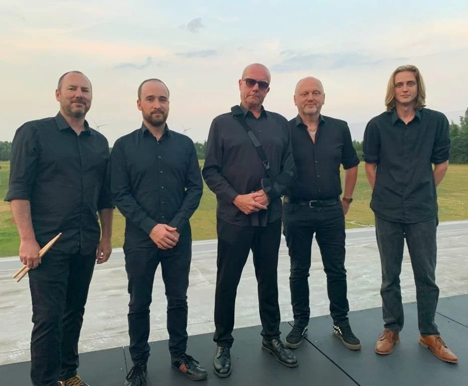 Vijf mannen staan buiten op een open veld, allemaal gekleed in zwarte kleding, met een bewolkte lucht op de achtergrond.