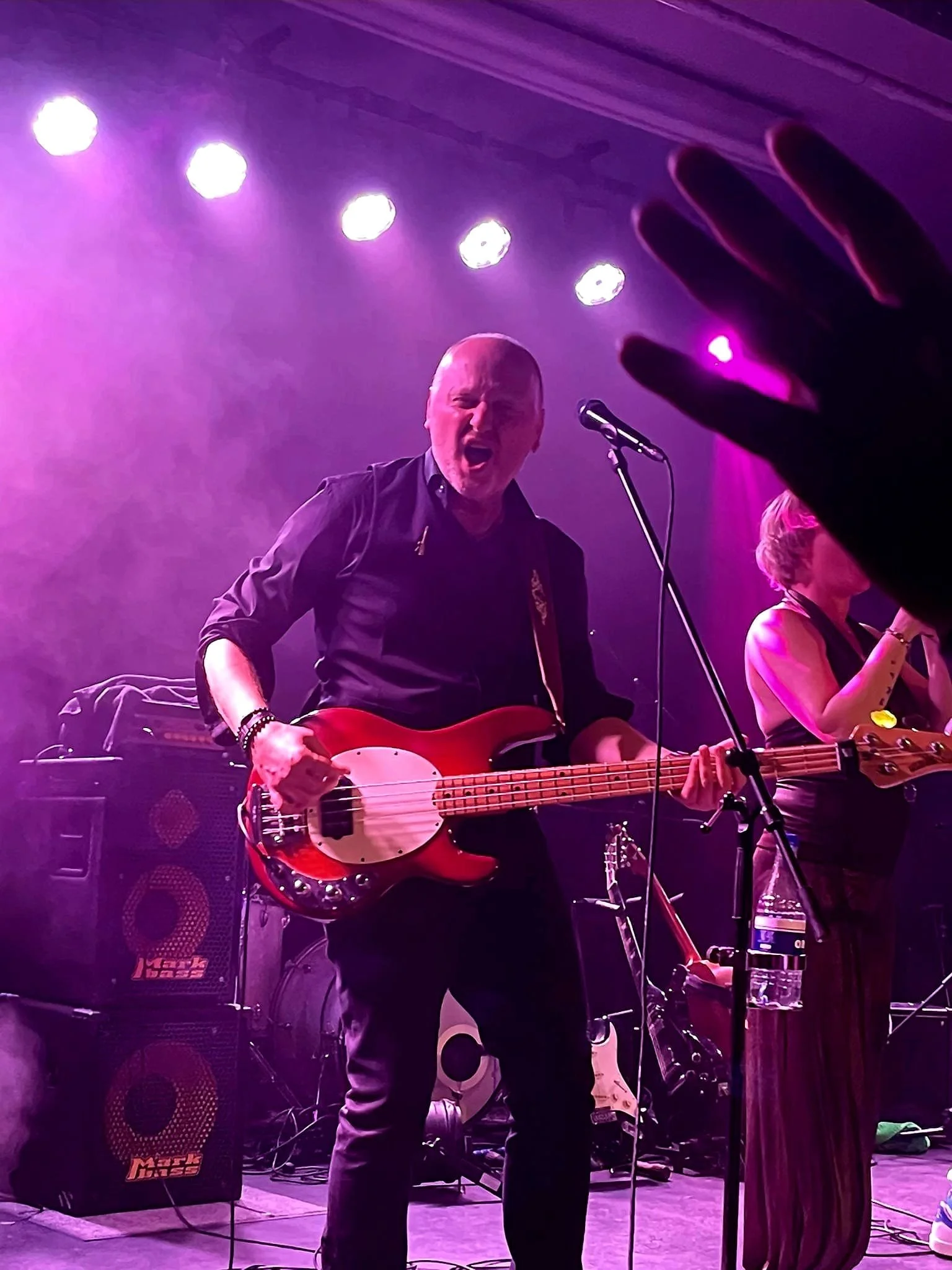 Muzikant speelt op een rode basgitaar tijdens een concert, omringd door muziekapparatuur en verlicht door roze en paarse podiumlichten.