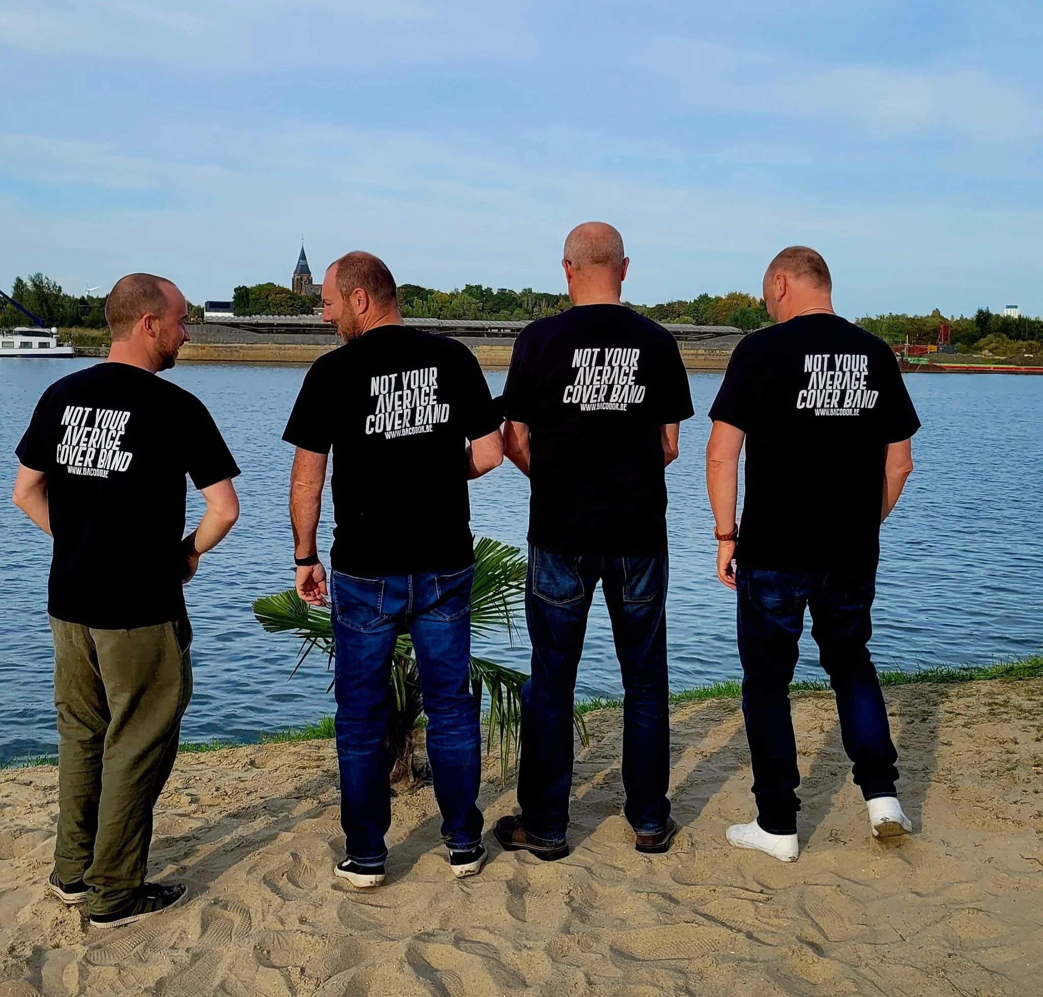 Vijf mannen staan aan de rand van een rivier, gekleed in zwarte T-shirts met de tekst 'NOT YOUR AVERAGE COVER BAND' op de achterkant. Ze kijken uit over het water, op de achtergrond zijn bomen, een kerk en gebouwen zichtbaar.
