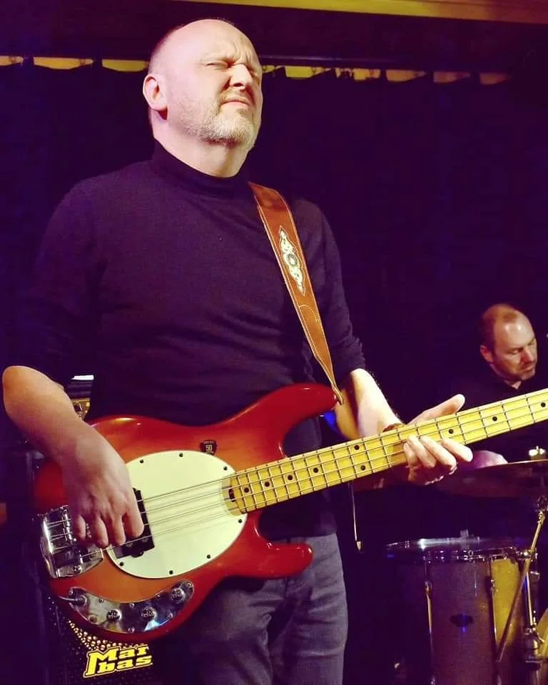 Man speelt basgitaar op een podium, met een andere muzikant op de achtergrond.