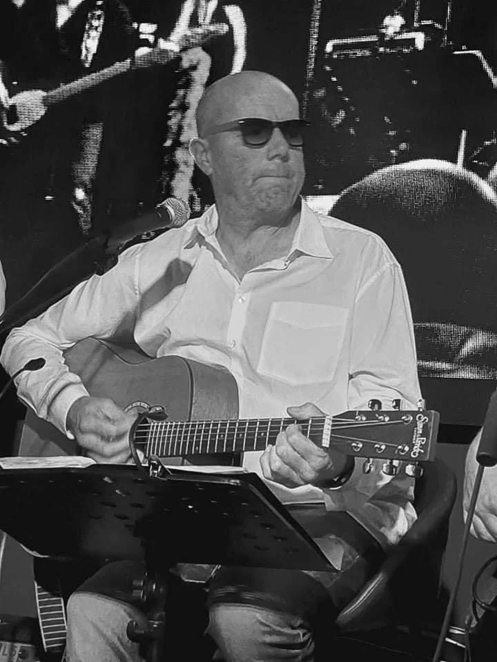 Man met een gitaar, draagt een wit overhemd en zonnebril, zit op een stoel, waarschijnlijk tijdens een muzikale sessie.