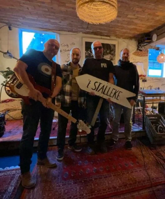Vier mannen staan binnen in een restaurant of café, één met een gitaar en een ander houdt een groot wit bord met de tekst '1 STALLEKE' vast.