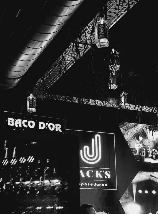 Een zwart-wit foto van een club of bar met neonlichten en enkele hangende lampen. Voorgrond met silhouet van een persoon en op de achtergrond neonborden met teksten zoals "BAC D'OR" en "JACK'S".
