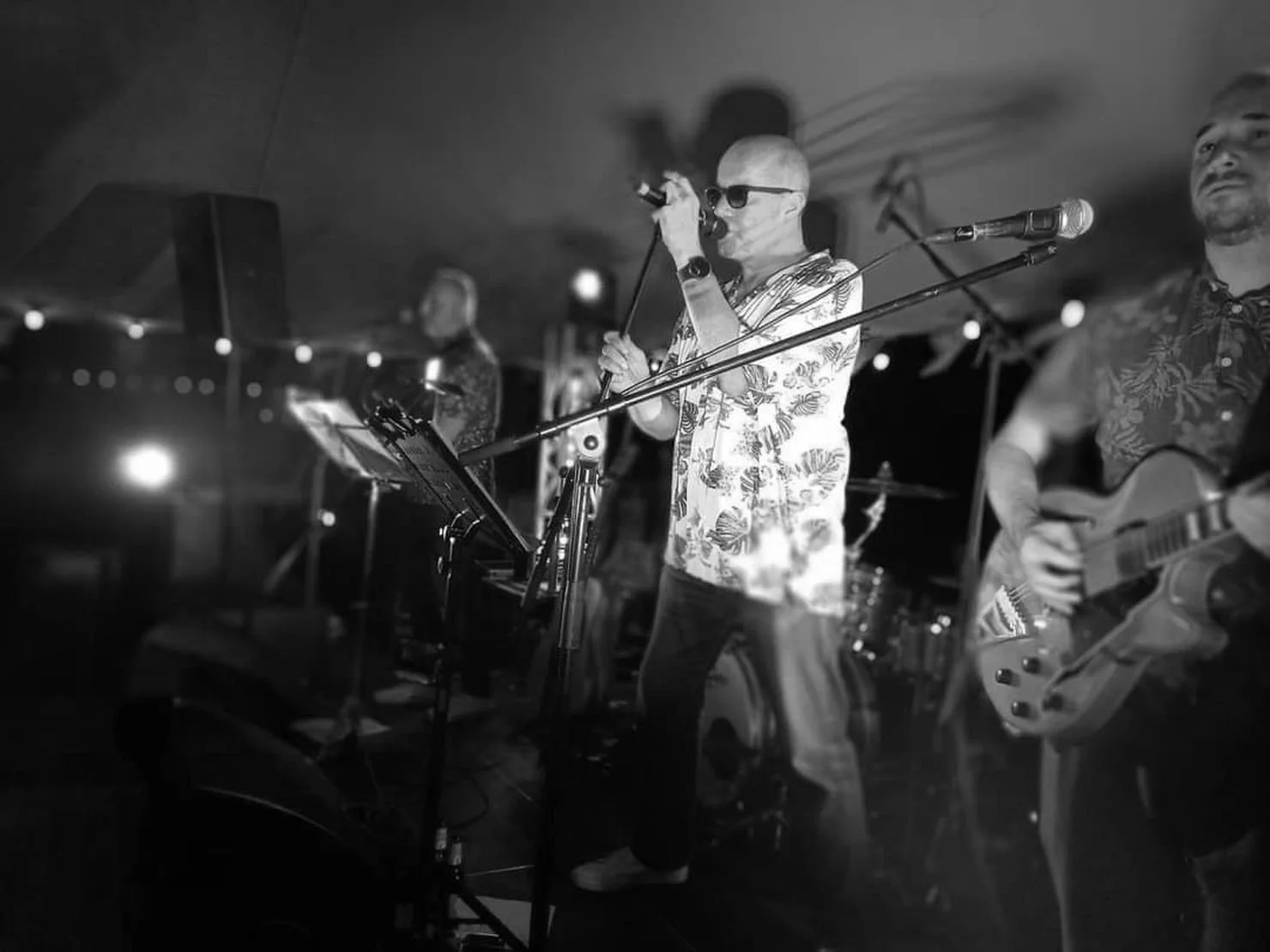 Mannen met een zonnebril zingt in een microfoon, samen met een gitarist en andere bandleden op een podium, op een donkere feestlocatie.