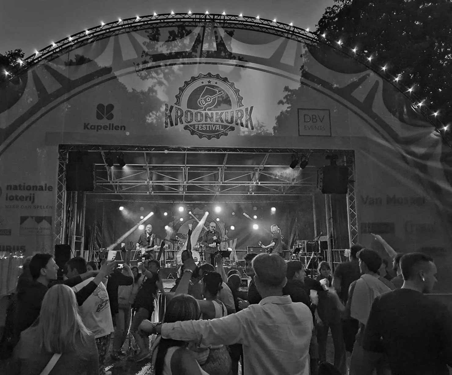 Een zwart-wit foto van een muziekfestival met een podium waarop een band speelt. Het publiek staat voor het podium en geniet van de optredens, sommigen maken foto's of bewegen mee met de muziek.