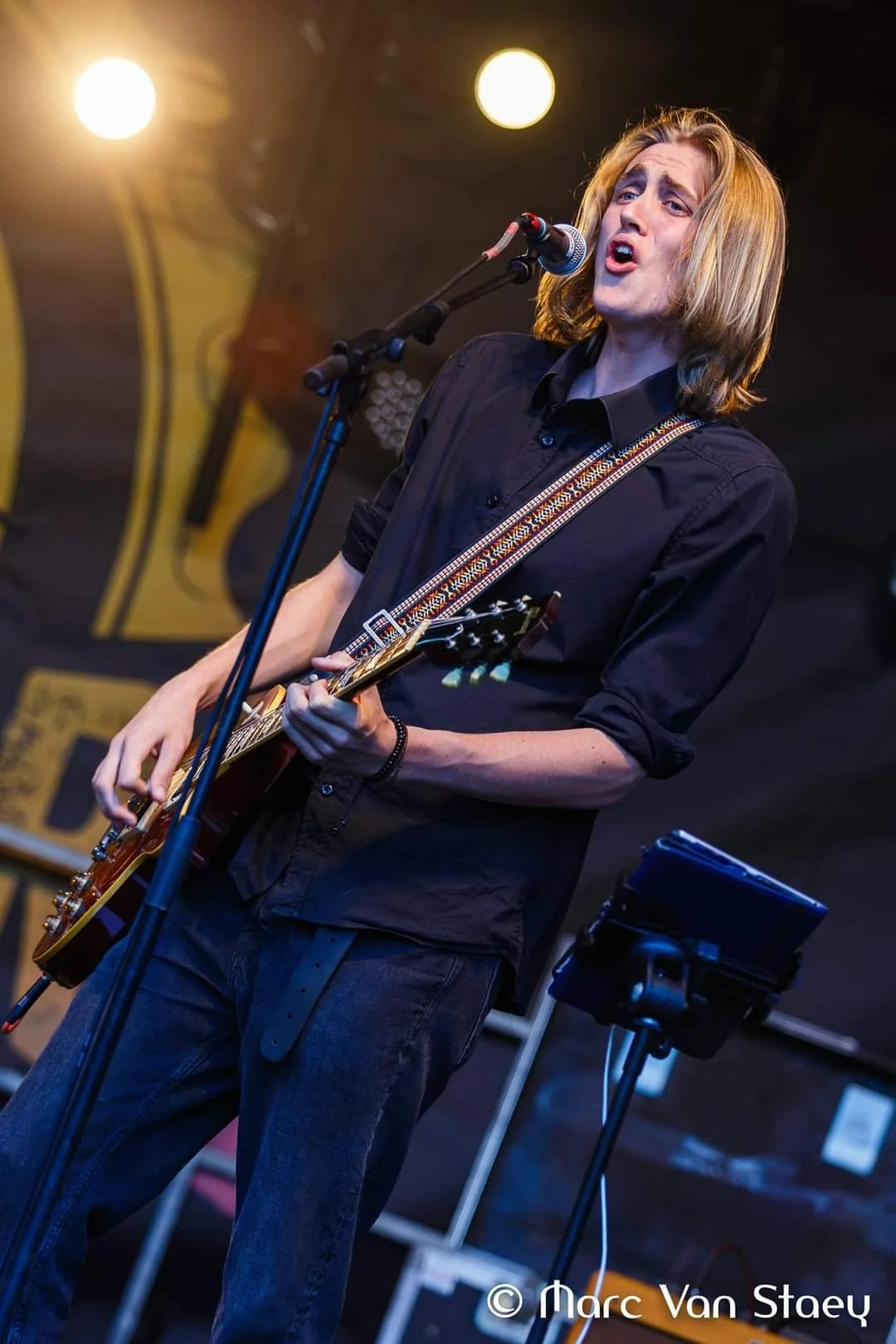 Mannelijke muzikant met lang blond haar speelt gitaar en zingt voor een microfoon tijdens een optreden. Op de achtergrond een licht en een affiche.