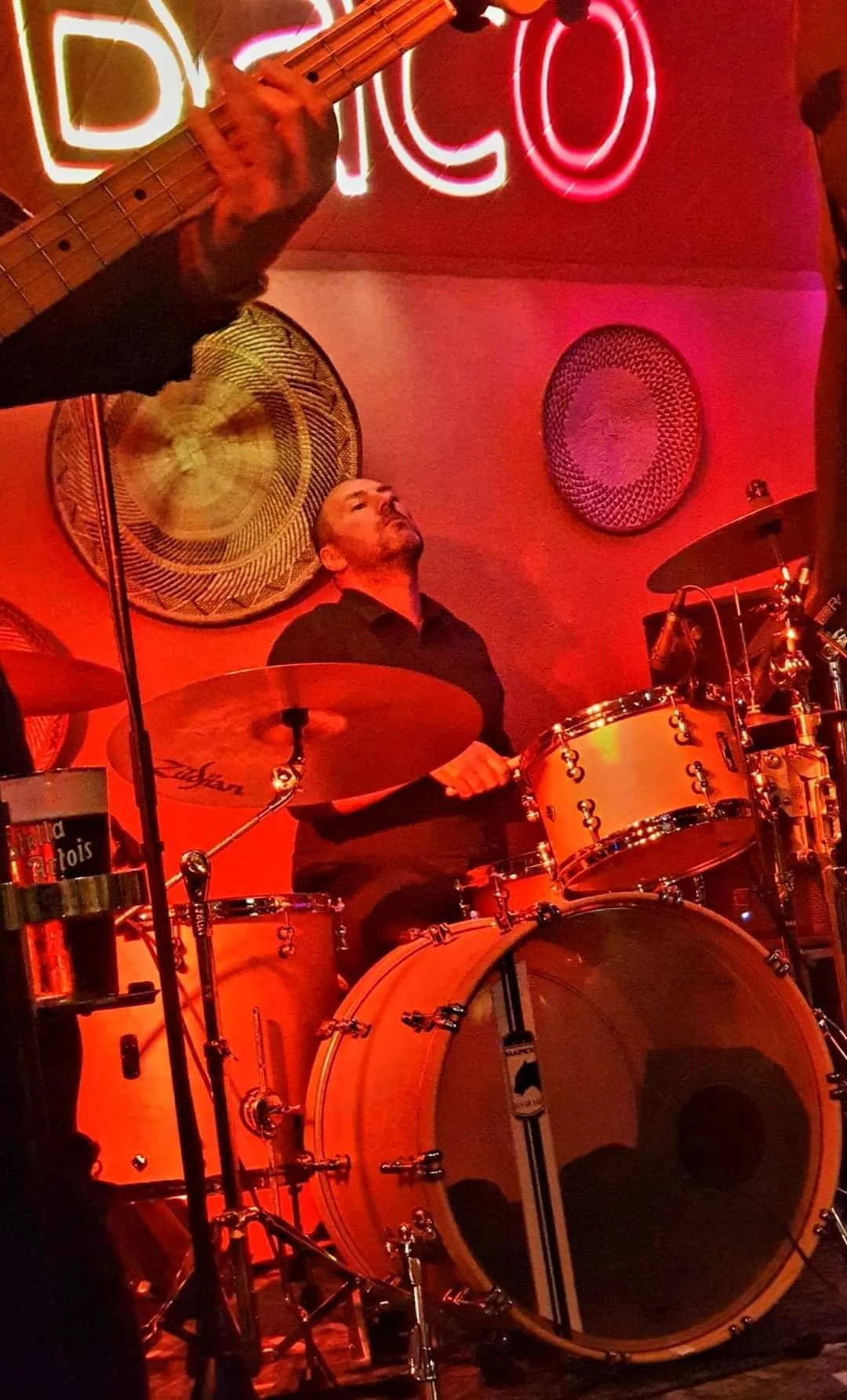 Een man die op een drumstel speelt in een donkere, gezellige gelegenheid met rode verlichting en neonborden.