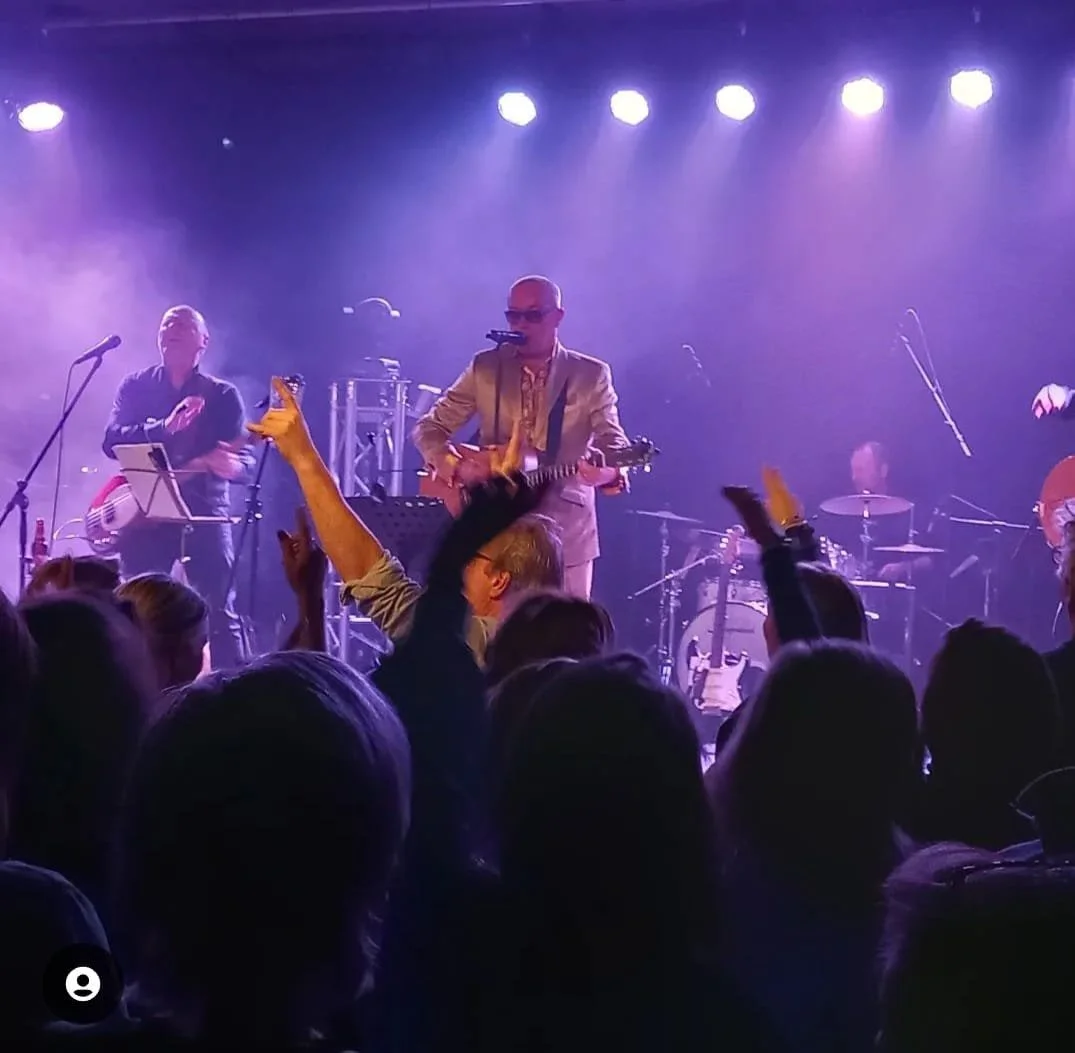 Een live muzikale uitvoering op een donker podium met blauwe en paarse verlichting, muziekinstrumenten en een zanger met een gitaar, omringd door een publiek dat juicht en de handen in de lucht steekt.