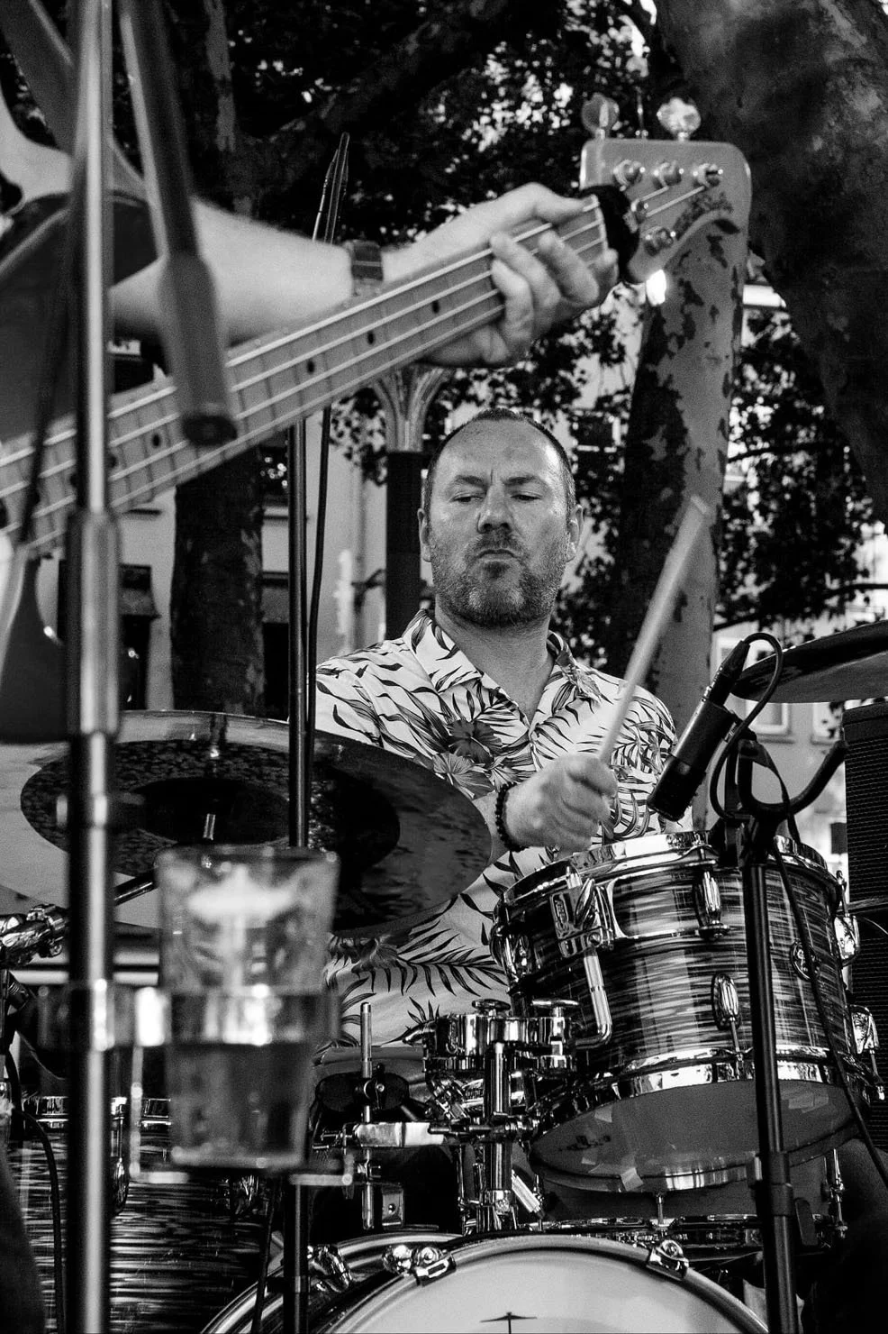 Een man die drums speelt tijdens een optreden, met een gitaarvast in de achtergrond, alles in zwart-wit.