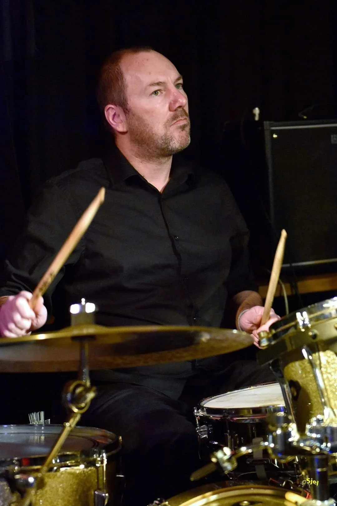 Man zit achter een goldkleurige drumset, draagt een zwarte overhemd en houdt drumstokjes vast, bezig met muziek maken.