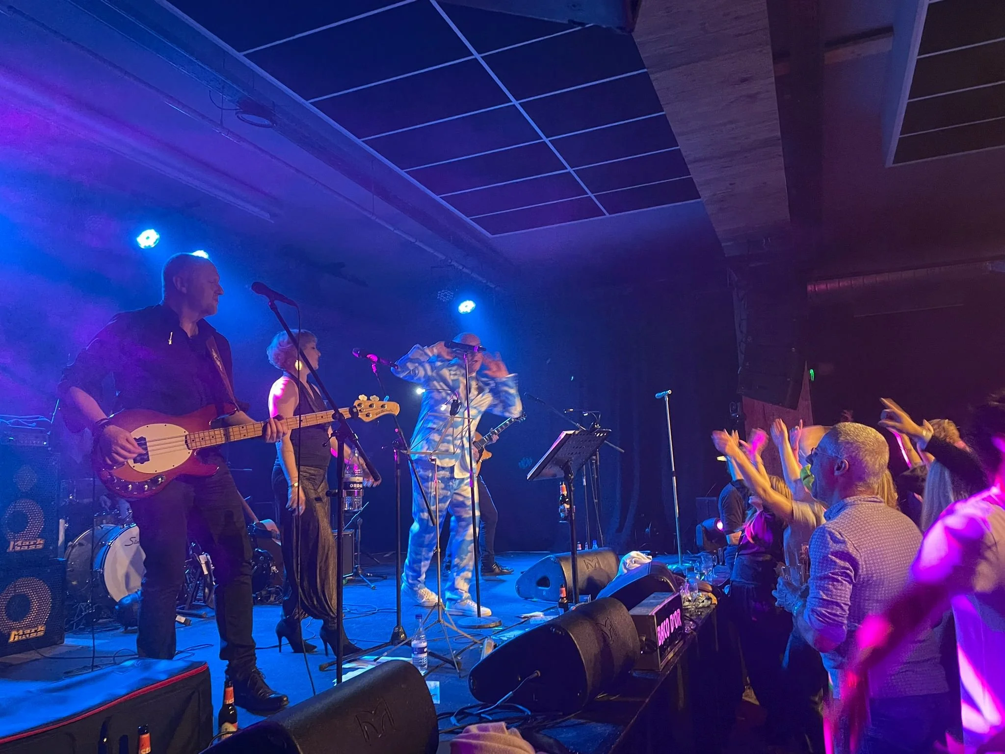 Een live muziekoptreden met een band op een podium en een publiek dat danst en plezier heeft.