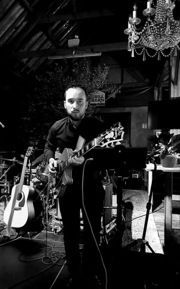 Man met een gitaar op een podium, omringd door andere muziekinstrumenten en apparatuur, binnen een sfeervolle avondsetting met kroonluchter.
