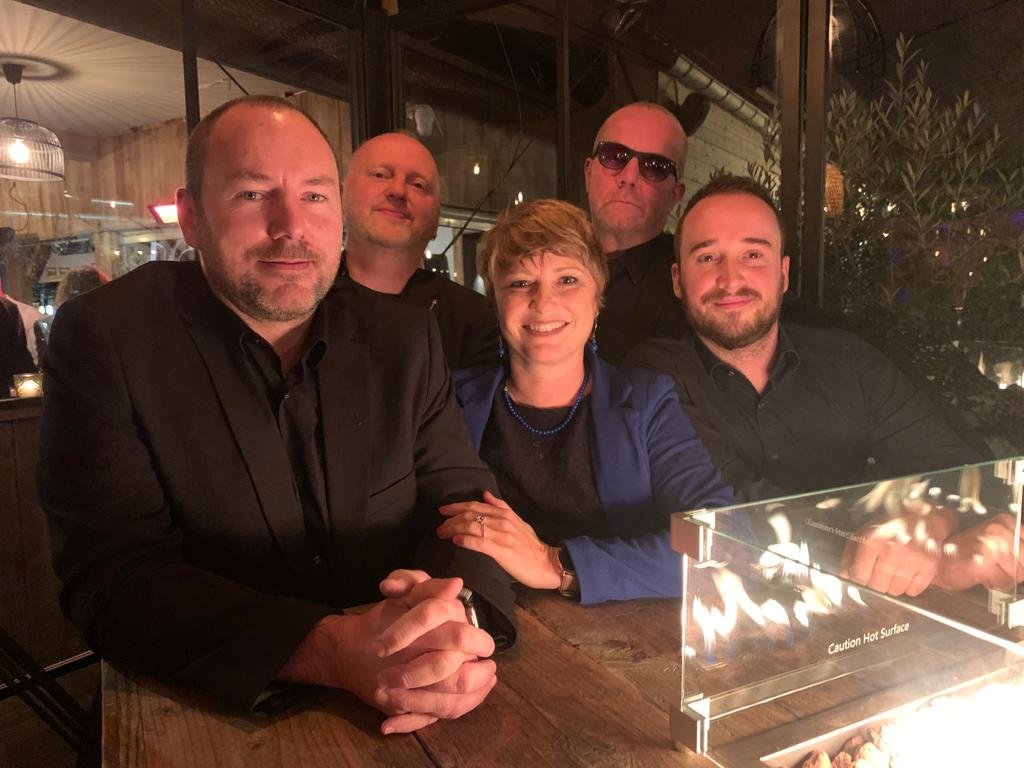 Vijf mensen zitten samen aan een tafel in een restaurant, met een vuurkorf op tafel, in een gezellige binnenruimte of terras, 'Caution Hot Surface' tekst zichtbaar op de vuurkorf.