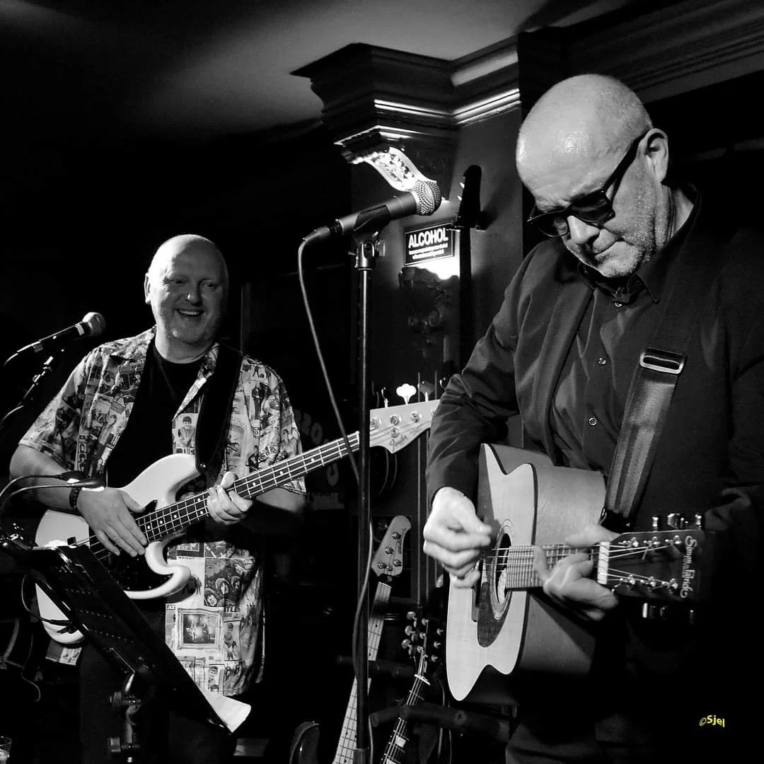Twee mannen die muziek maken met gitaren in een rokerige, gezellige ruimte, zwart-wit foto.
