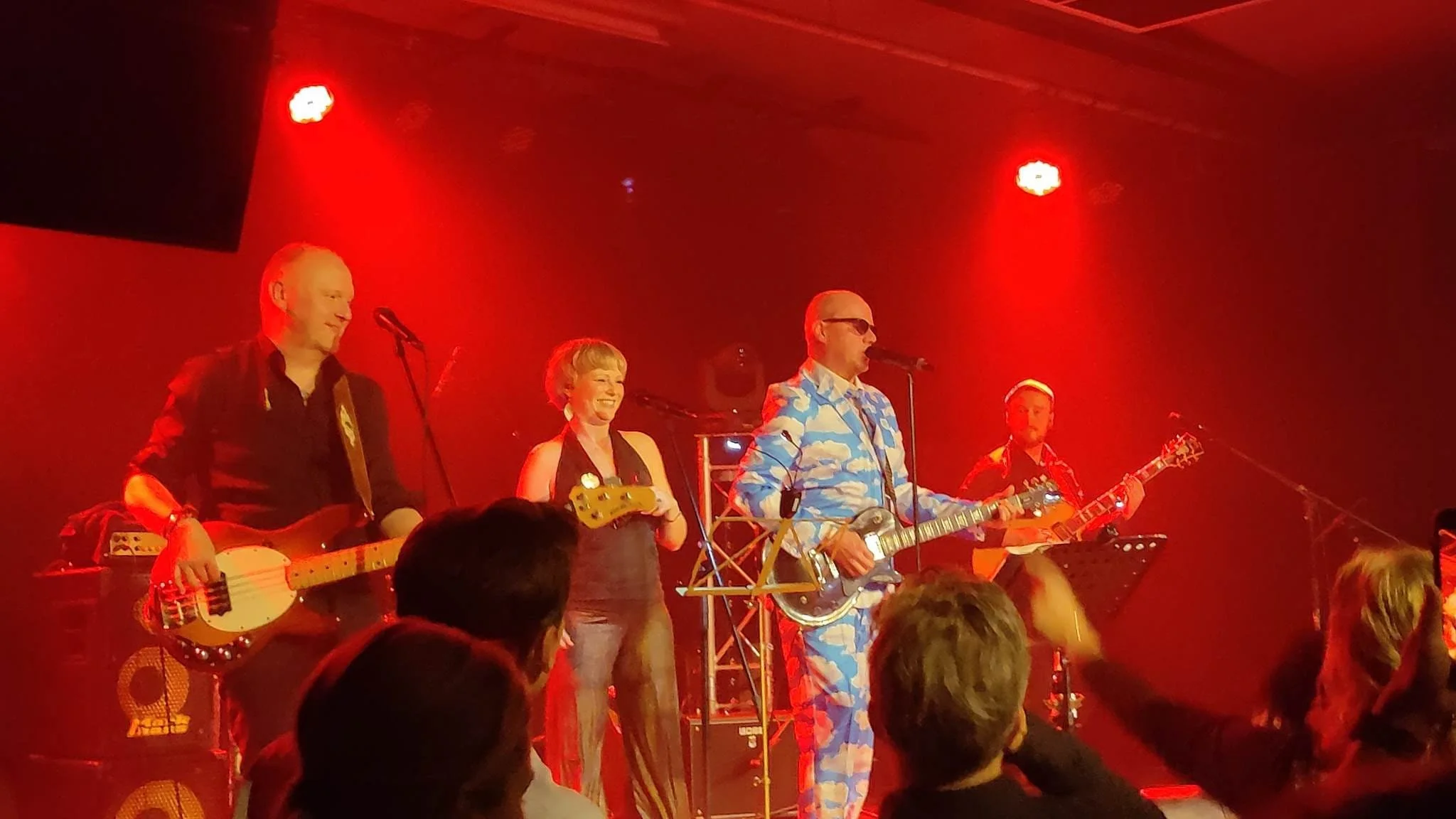 Mannen en vrouw op een podium met gitaren en microfoons, optreden onder rode verlichting, publiek zichtbaar op voorgrond.