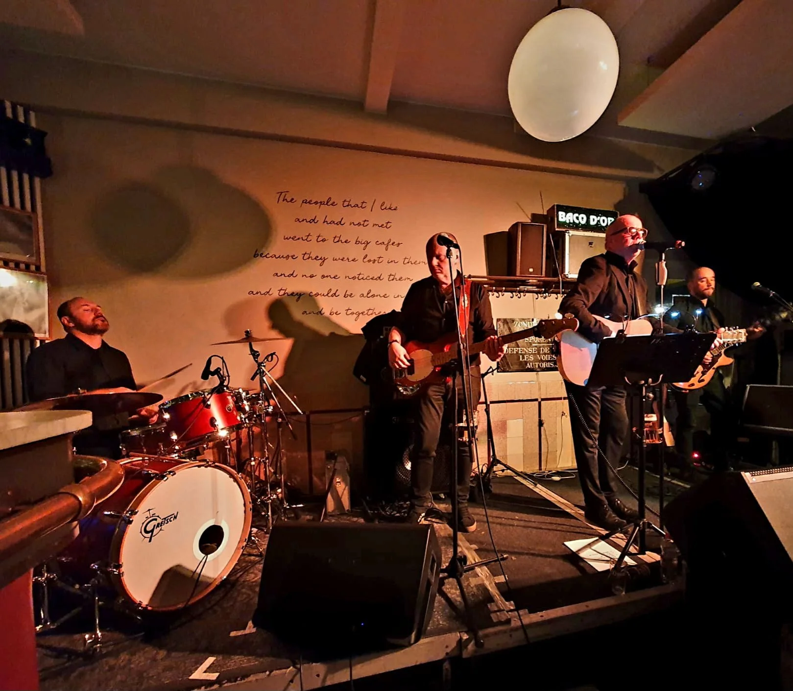 Een band speelt live muziek op een podium in een club, met een drummer, twee gitaristen en een zanger, onder warme verlichting.