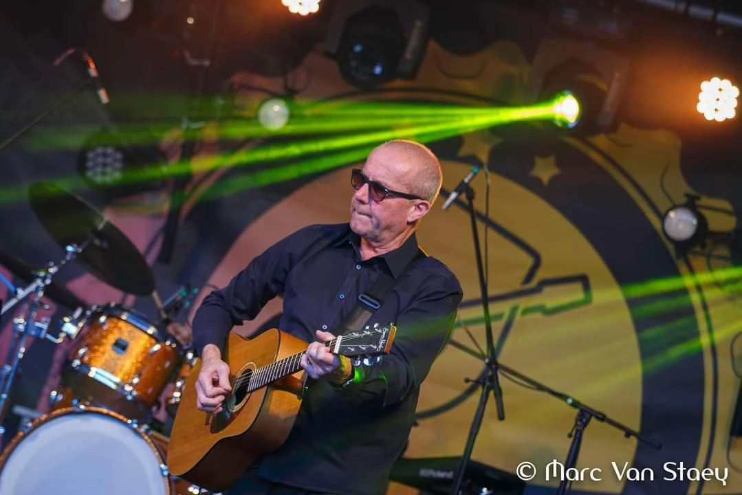 Mannelijke performer met een gitaar op een podium met kleurrijke lichteffecten en een drumstel op de achtergrond.