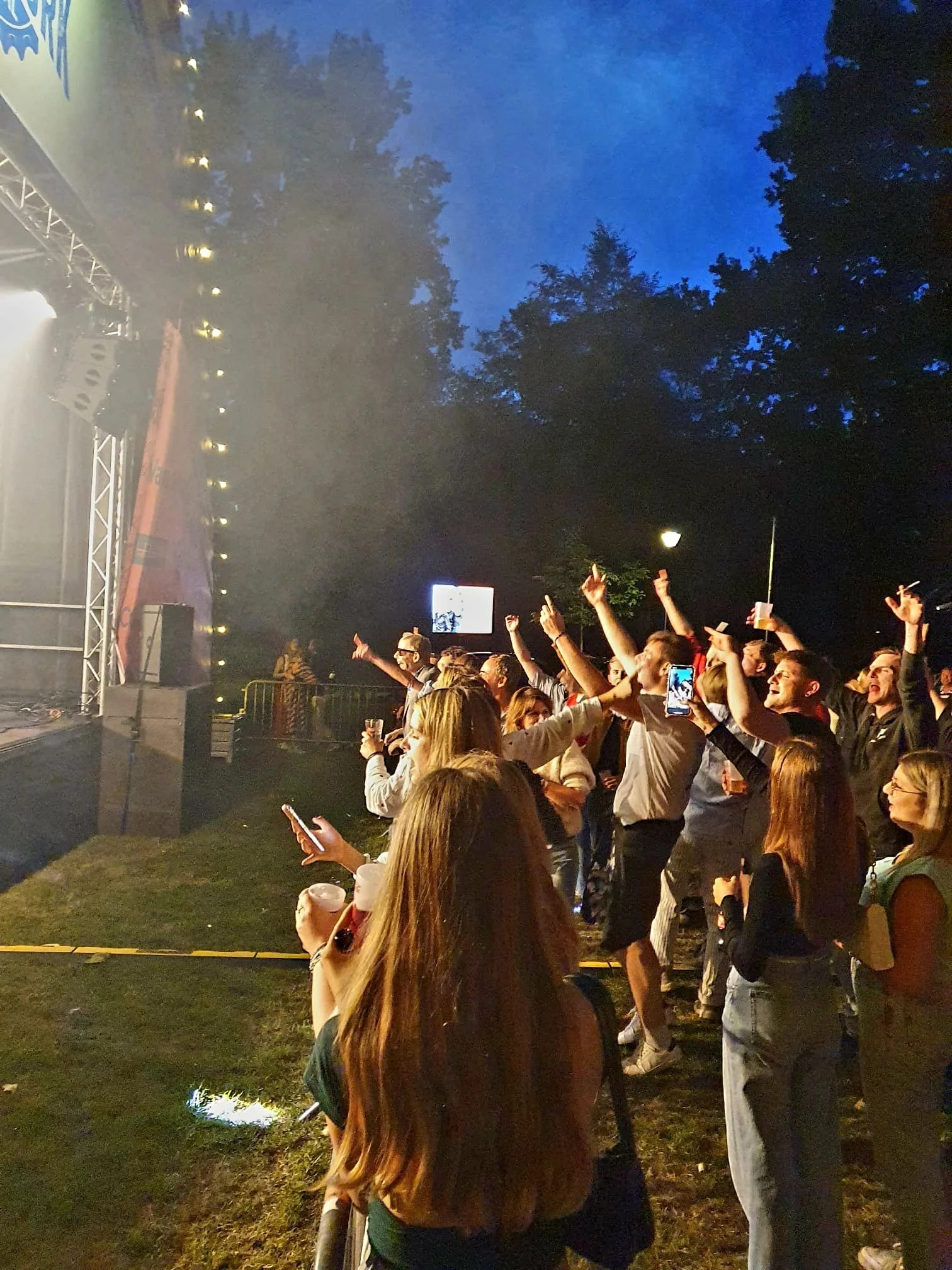 Mensen genieten van een openluchtconcert in de avond, dansend en photographend, met een verlicht podium en blauwe avondhemel.