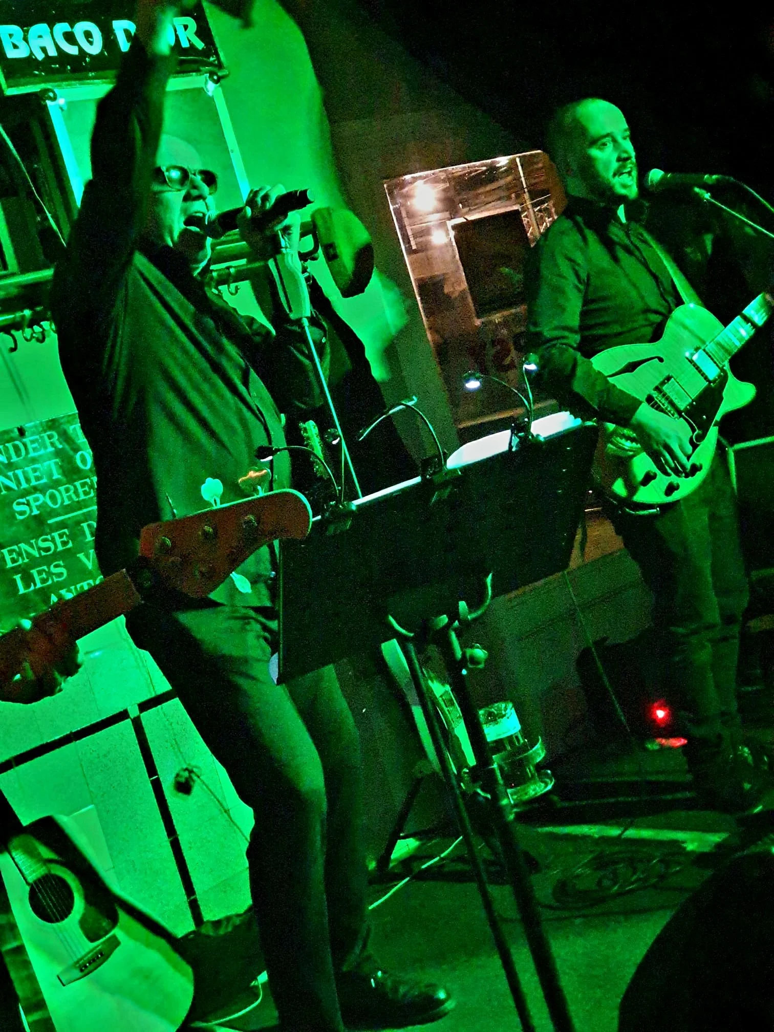Mensen die live muziek spelen op een podium in een bar, verlicht door groene verlichting, met gitaren en microfoons.