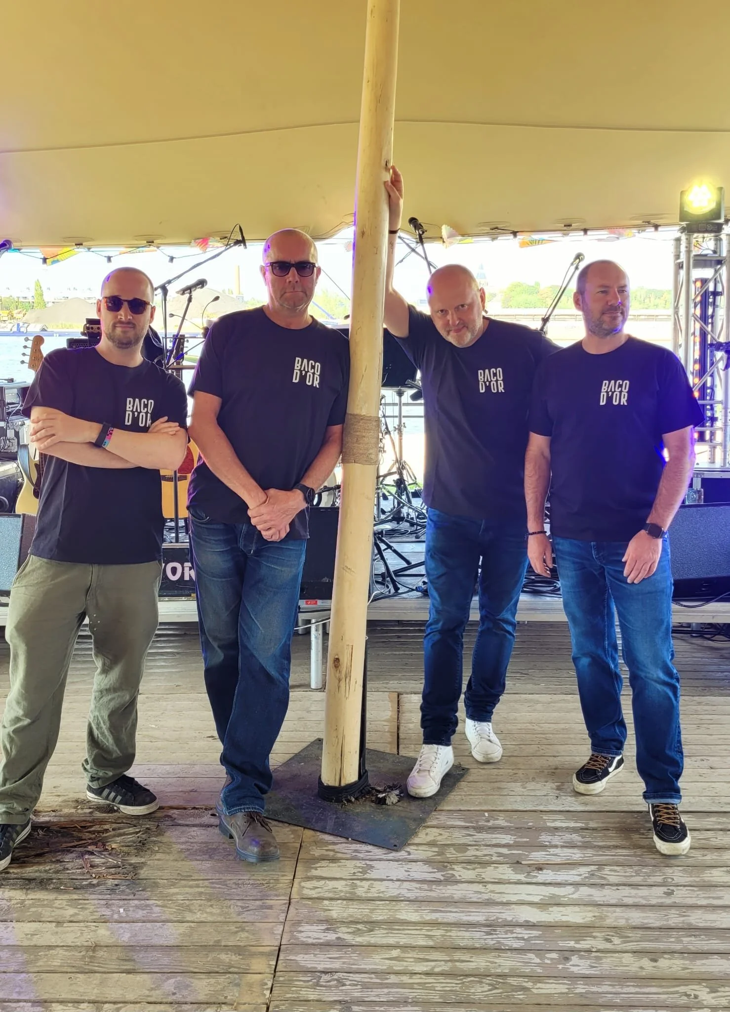 Vier mannen staan onder een grote tent met muziekapparatuur op de achtergrond, allemaal dragen ze een zwart t-shirt met de tekst 'BAC D'OR'.