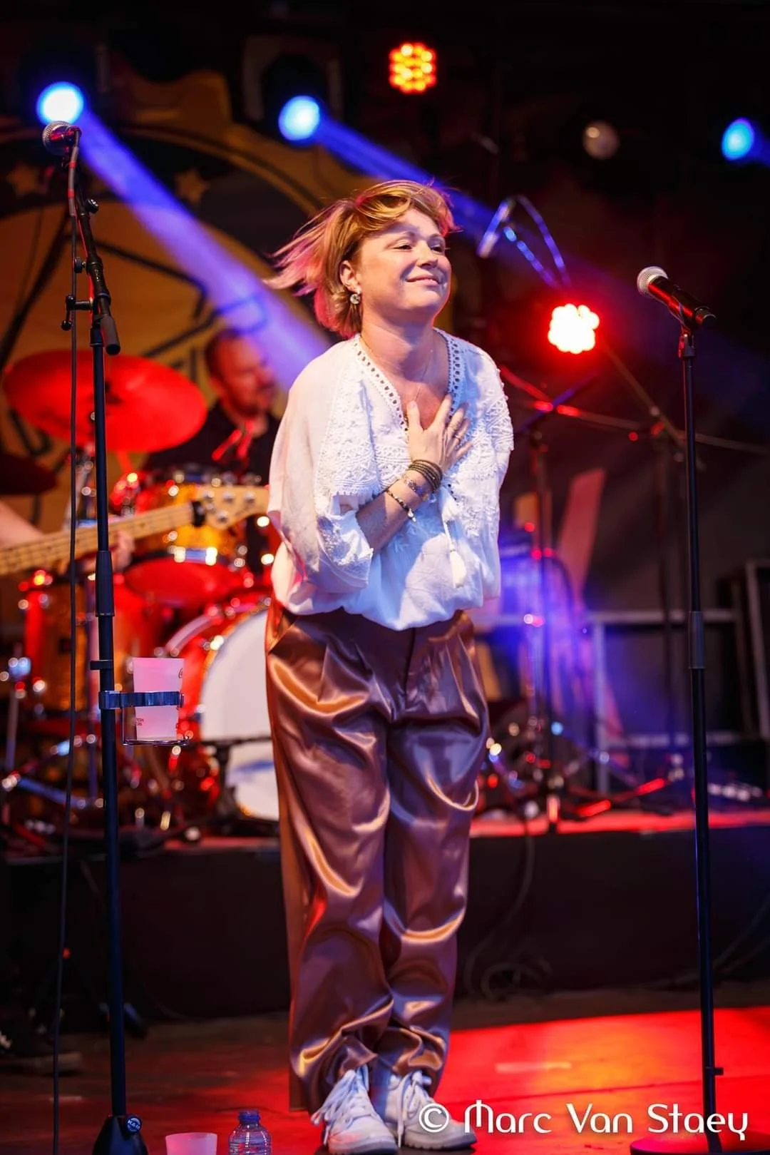 Een vrouw staat op een podium met haar hand op haar borst, terwijl ze naar het publiek kijkt. Achter haar speelt een band met een drummer. Er zijn kleurrijke podiumlichten, waaronder blauw en rood.