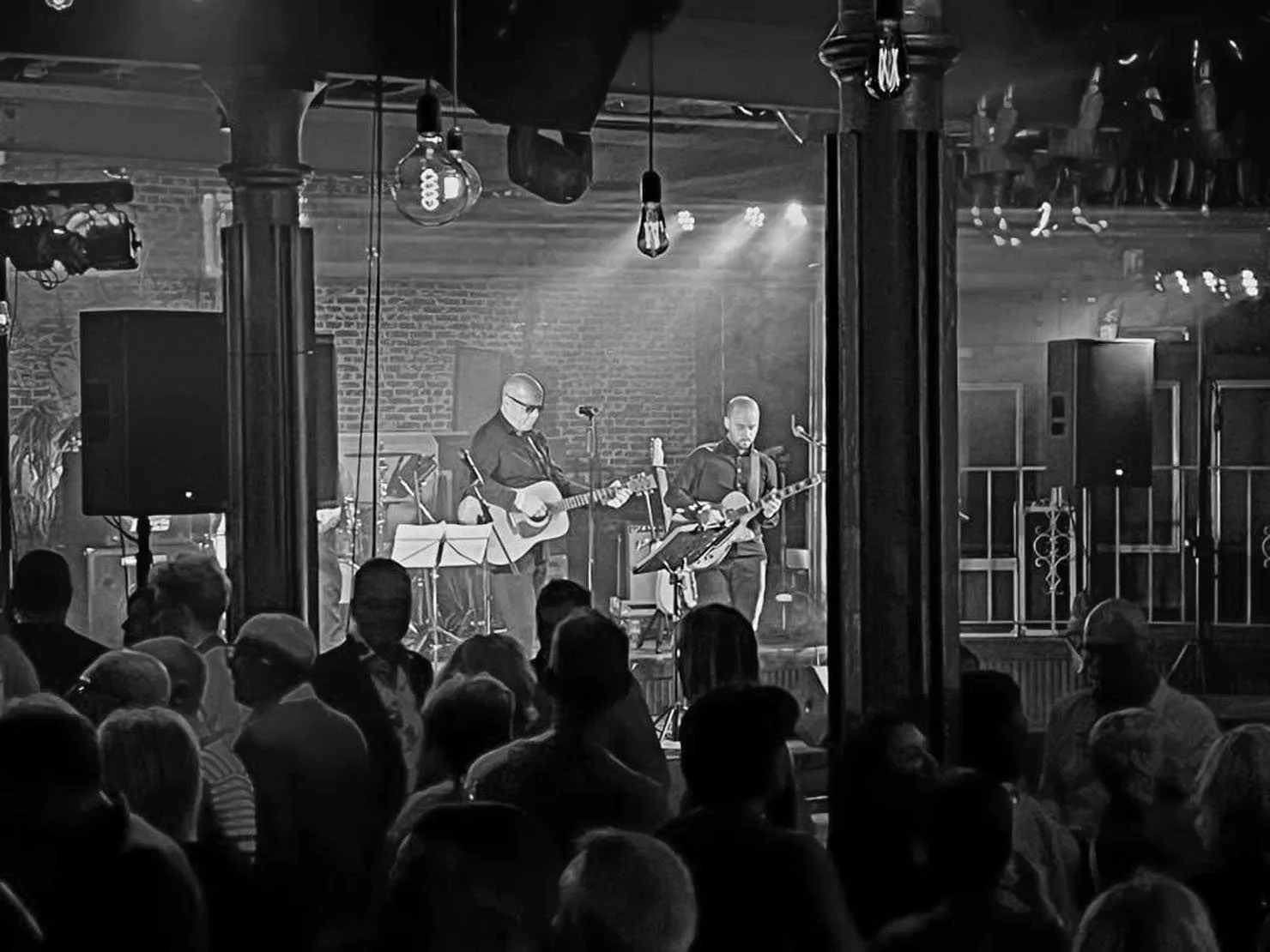 Een zwart-wit foto van een live muziekevenement met een band op een podium, twee gitaristen en een zanger, met een publiek dat naar de show kijkt.