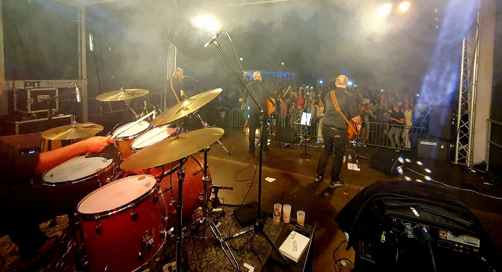 Muziekband speelt op een podium met een publiek dat danst en geniet, er is rook of mist en kleurrijk podiumlicht.