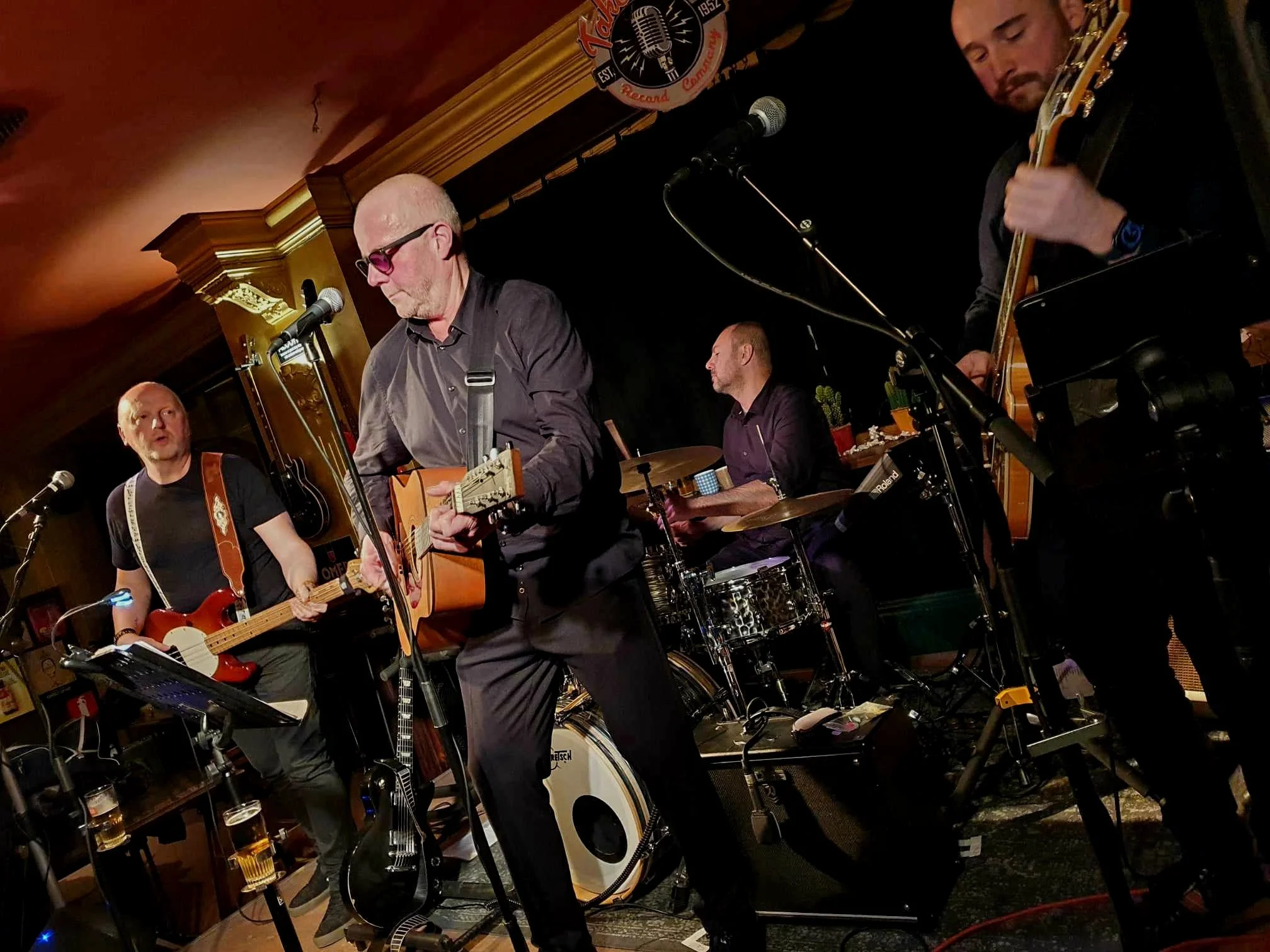 Mannen spelen muziek samen op een live podium in een club of café, met gitaren en drums, onder een plafond met gouden décor.