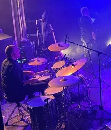 Drummer op een podium met een drumstel en een zanger die in de achtergrond staat, verlicht door blauwe en paarse lichten.