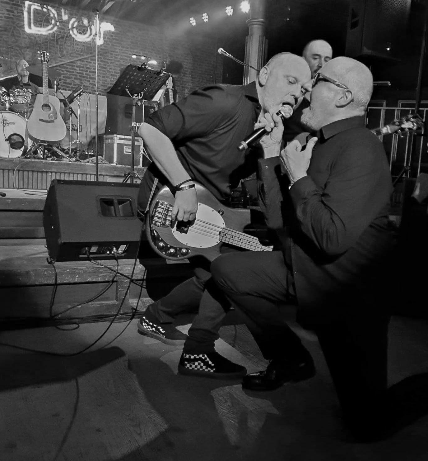 Twee mannen op een podium, één met een basgitaar, lijken te zingen of praten met elkaar, terwijl een band met gitaren en drums op de achtergrond speelt, in een zwarte en witte foto.