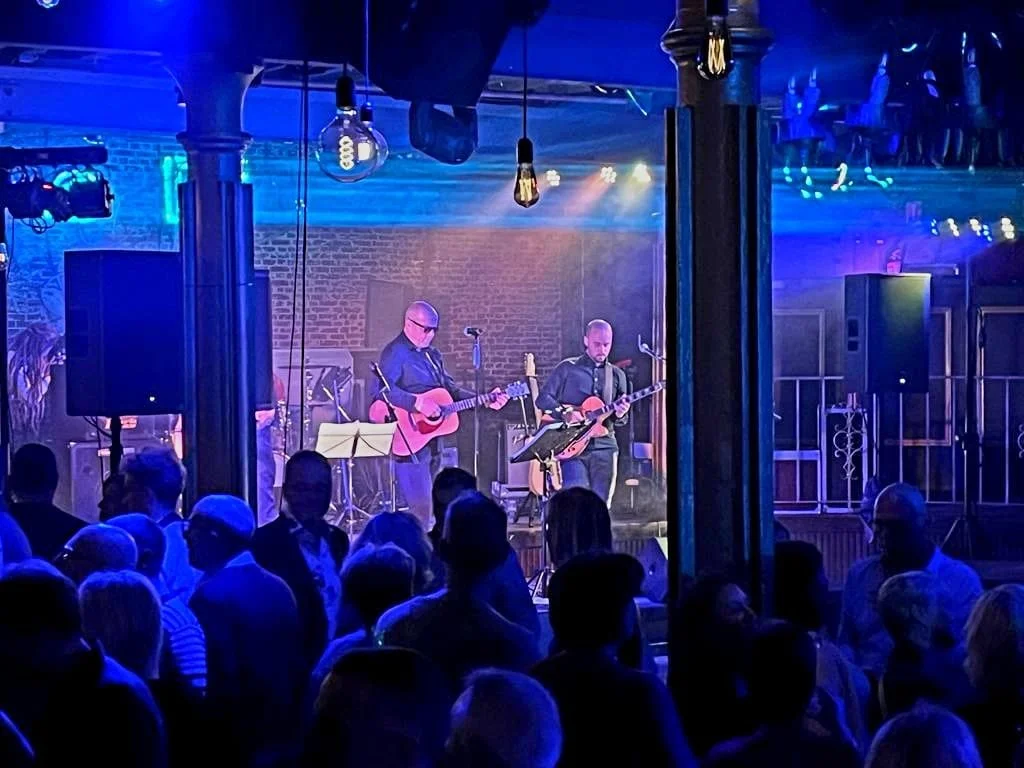 Twee muzikanten spelen op een podium met gitaren tijdens een live optreden, terwijl het publiek kijkt. Er is blauwe en paarse verlichting.