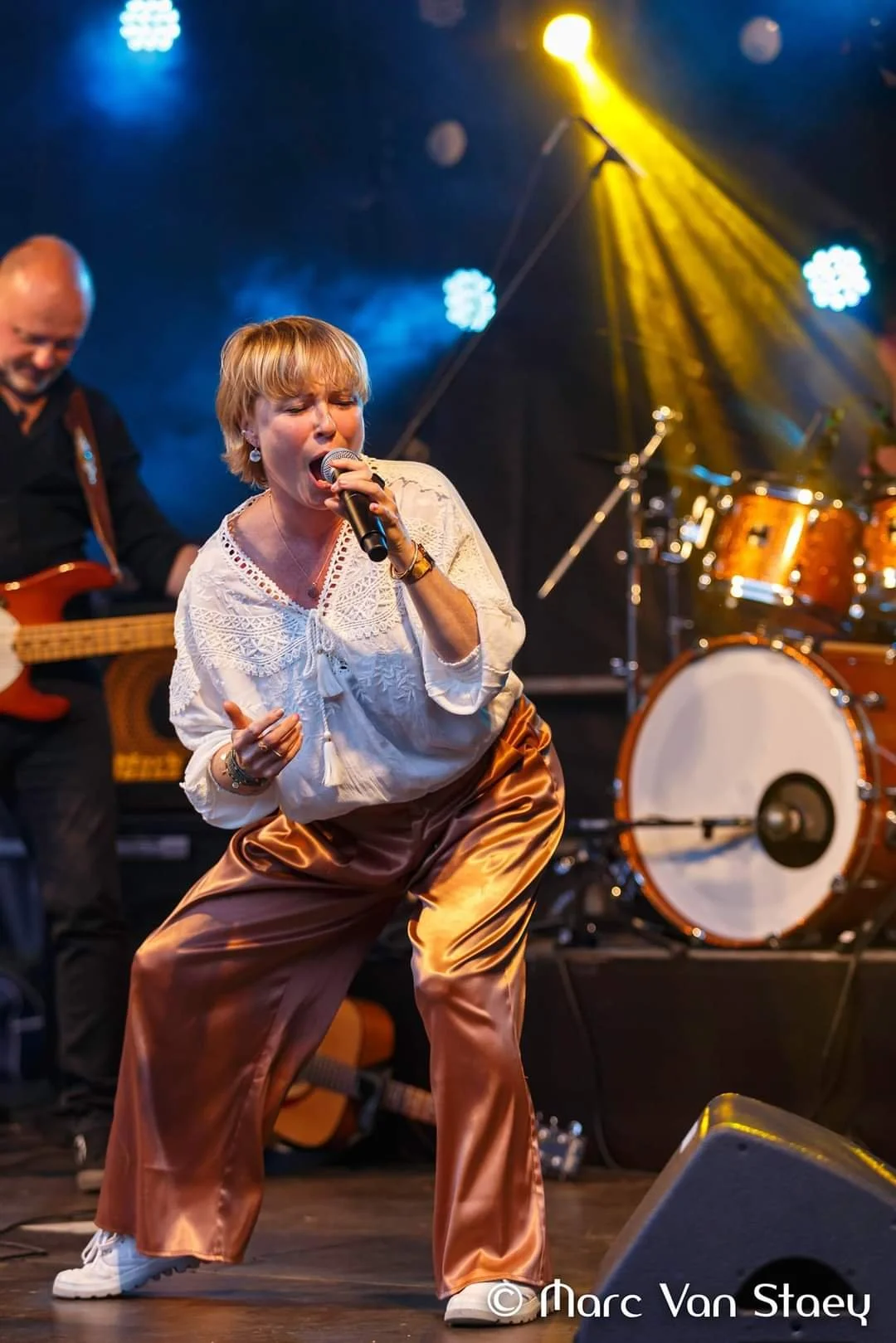 Een vrouw zingt in een band tijdens een optreden, met een man op de achtergrond die op een basgitaar speelt en een drumset op de achtergrond.