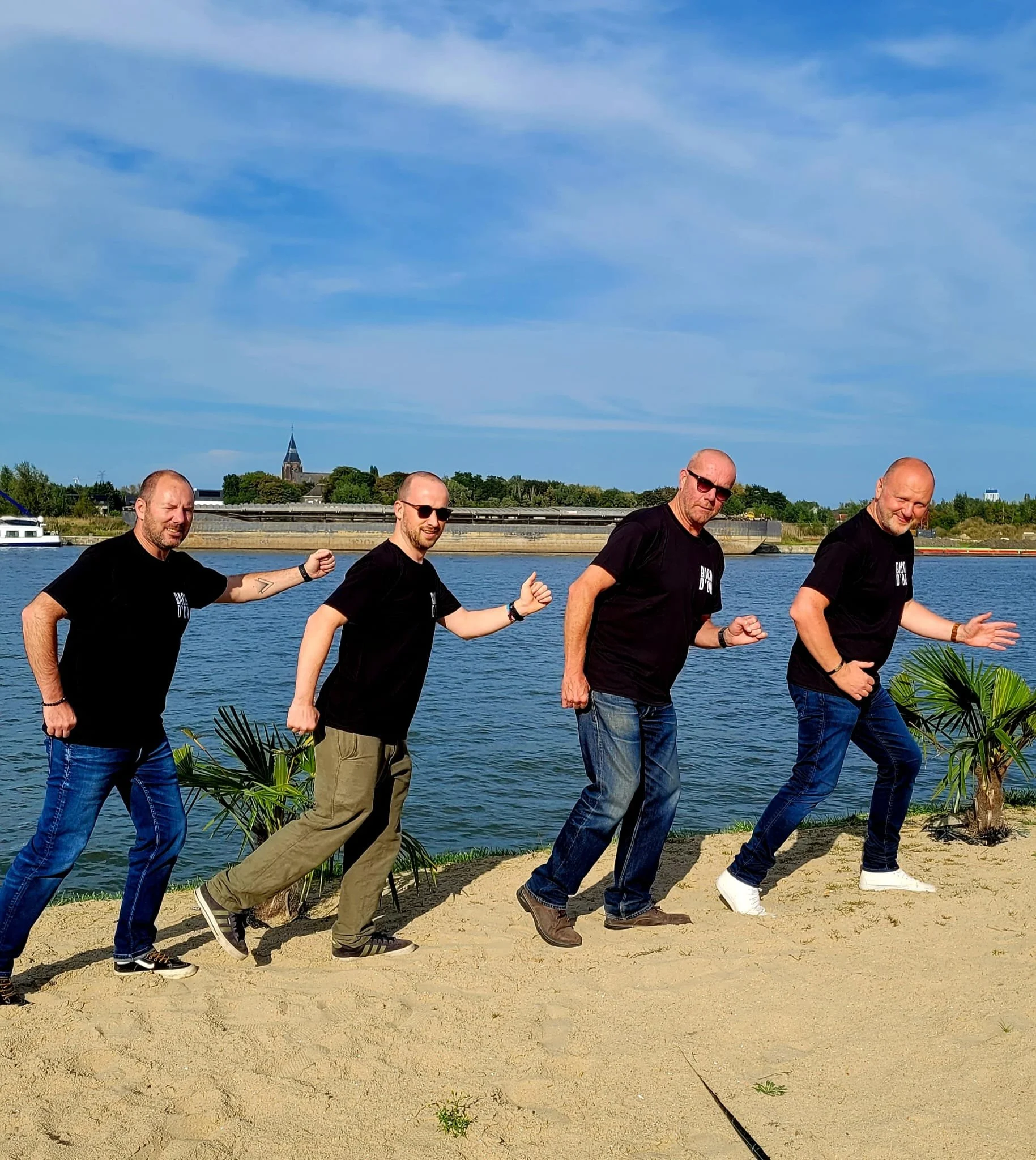 Vier mannen in zwarte t-shirts en jeans of khaki broek lopen op het zandstrand naast een rivier, met een blauwe lucht en een gebouw met een torenspits op de achtergrond.