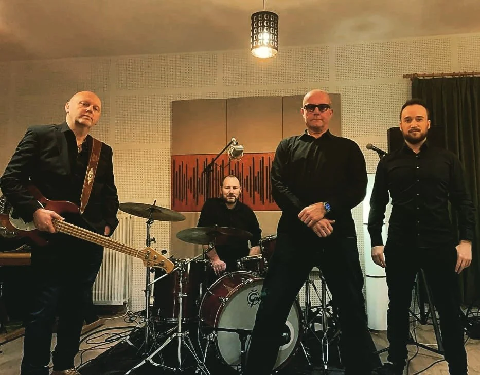 Vier mannen in zwart gekleed staan in een muziekstudio met instrumenten: een man met een basgitaar, een man achter de drums, een man met zonnebril en een man zonder zonnebril.