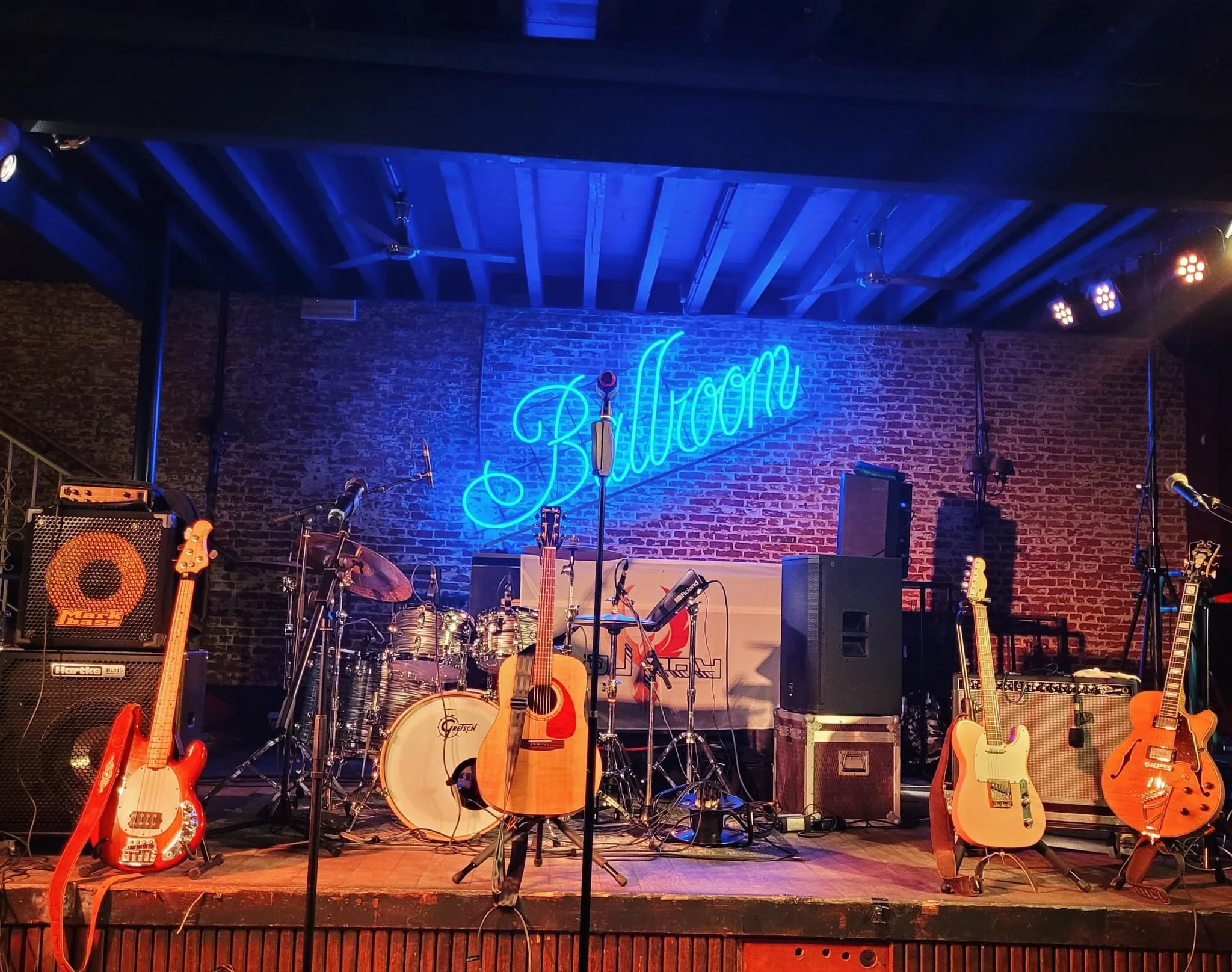 Muziekpodium met elektrische gitaren, akoestische gitaren, een drumstel, microfoons en versterkers, met een blauwe neonlichtsignaal dat 'Balloon' zegt op een bakstenen muur.