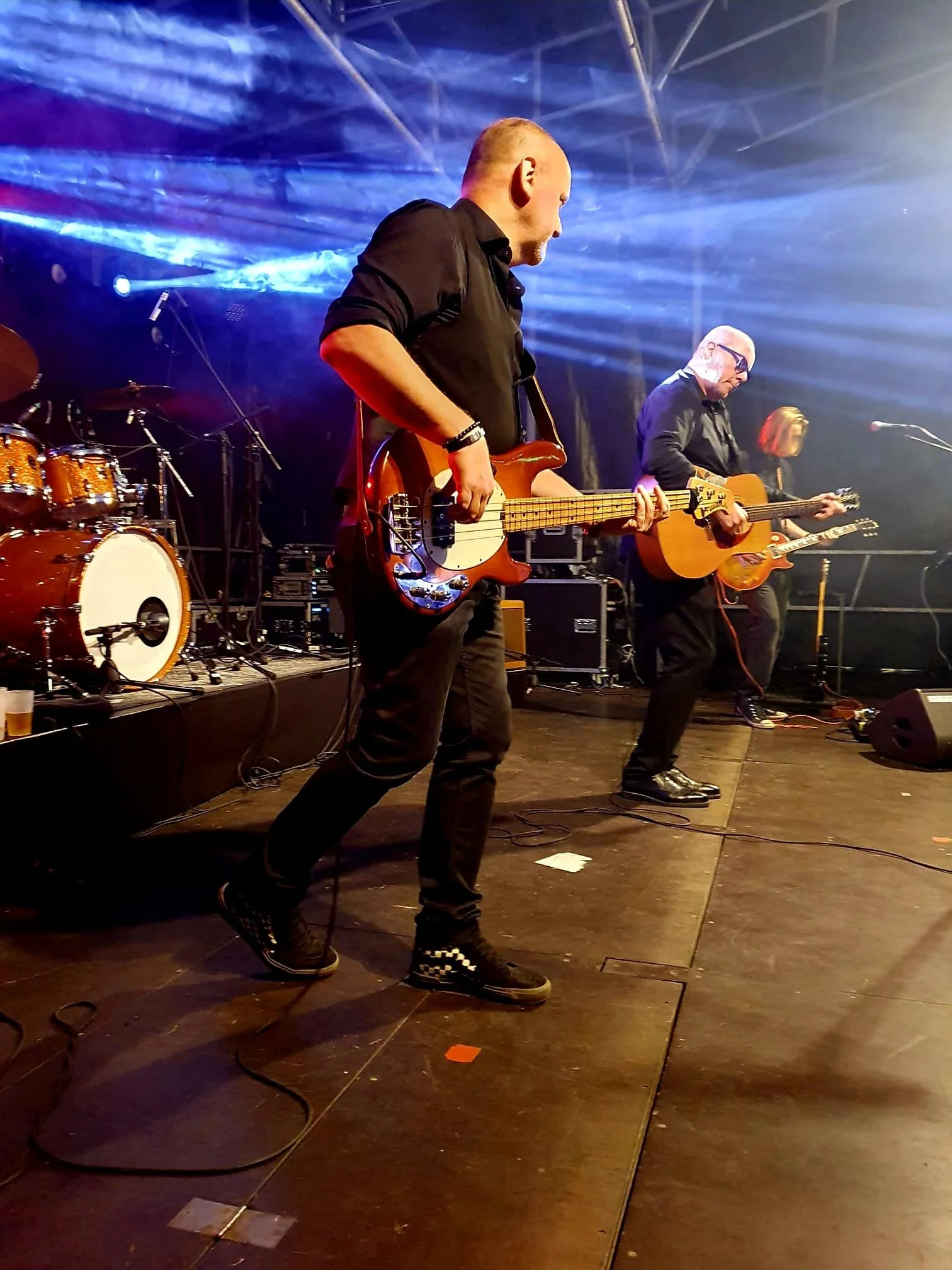 Band speelt op een podium met verschillende muzikanten, waaronder een bassist en twee gitaristen, onder blauwe podiumverlichting.