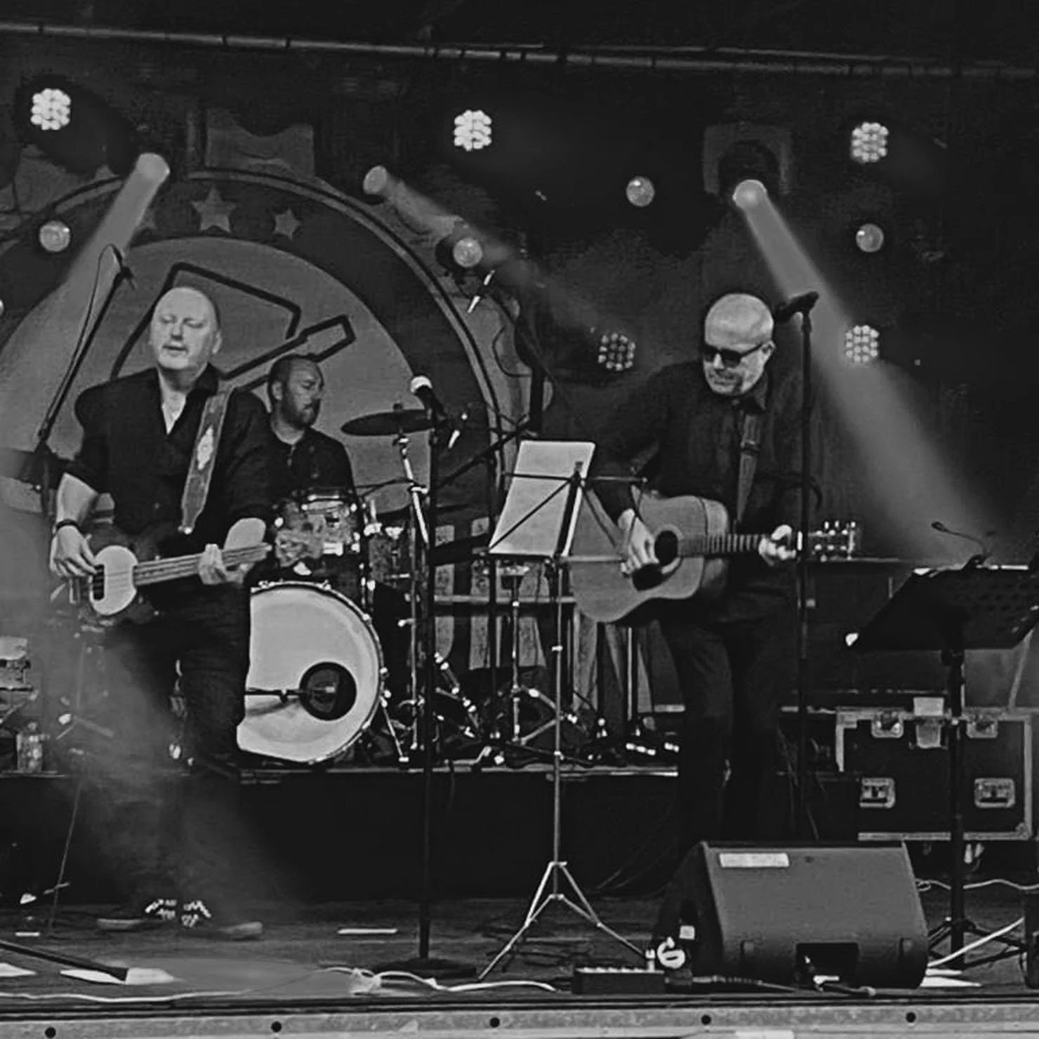 Muzikale band op een podium met drie leden: een bassist, een drummer en een gitarist, in zwart-wit.
