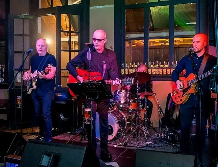 Vier muzikanten spelen op een podium met gitaar, drums en microfoons, binnen een gezellige, verlichte ruimte.