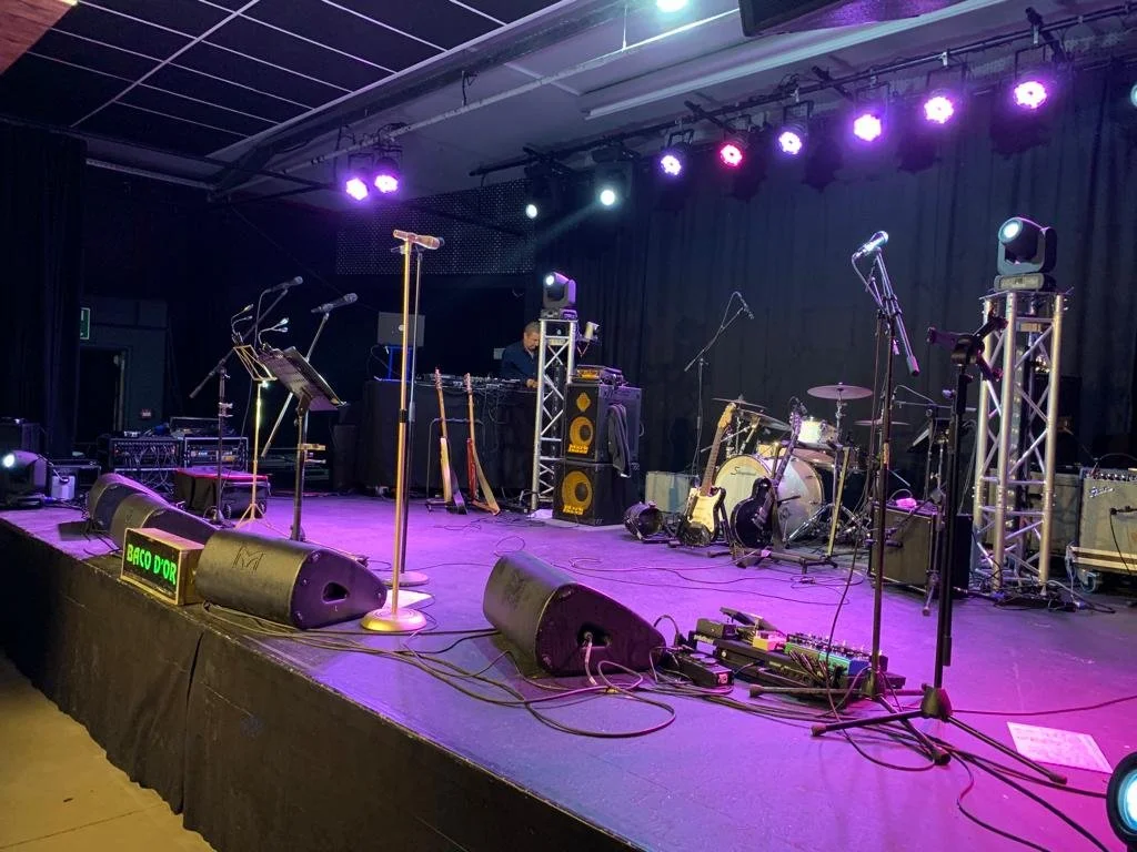 Podium met muziekinstrumenten en banduitrusting, klaar voor een optreden, met koperenoortjes en gitaren, onder kleurrijk podiumverlichting.