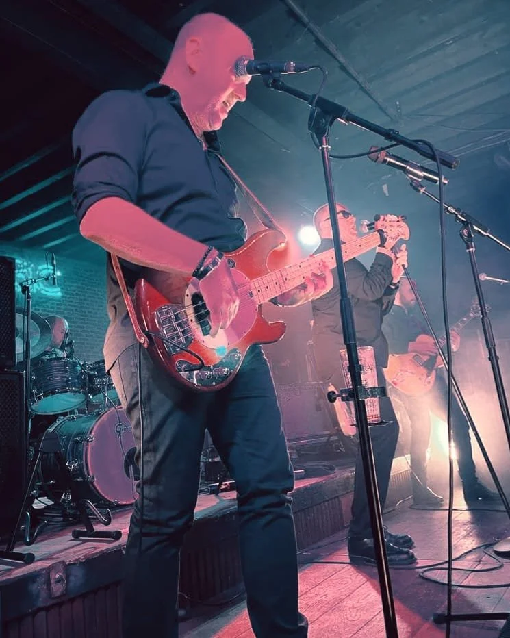 Mannen die live muziek speelt op een podium, met gitaren en een drumstel op de achtergrond, onder kleurrijk podiumlicht.