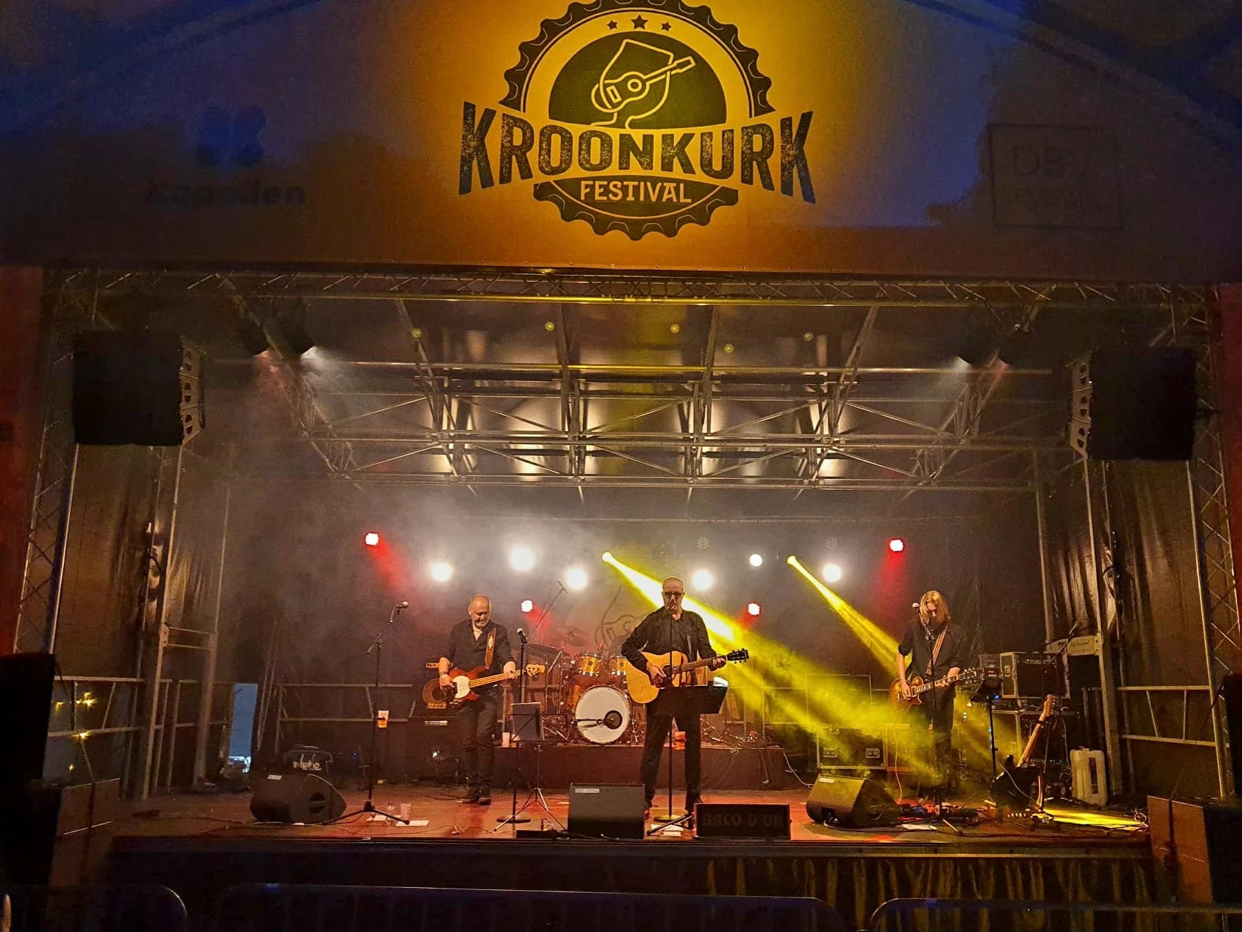 Podium met een band die live speelt tijdens Kroonkurk Festival, met drie muzikanten onder kleurrijke lichten, en een groot festivalbanner erboven.
