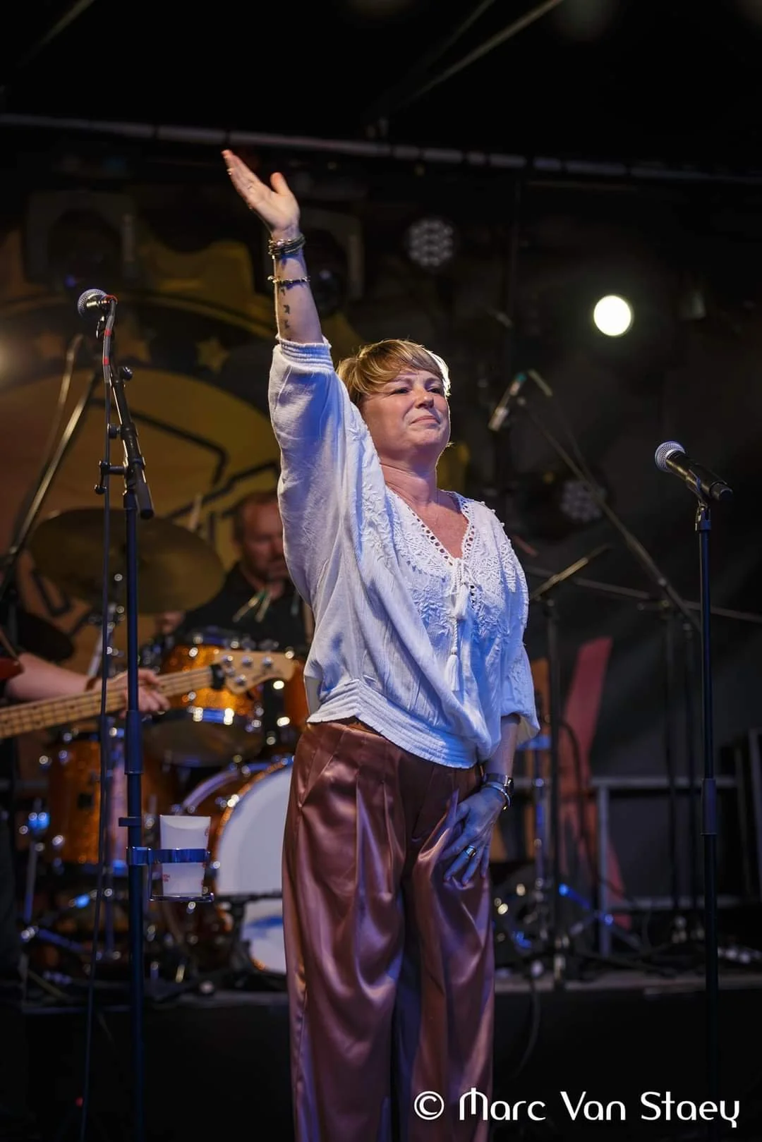 vrouw op het podium met hand omhoog, muzikanten op de achtergrond, tijdens een concert.