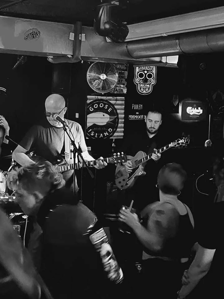 Een zwart-wit foto van een live muziekoptreden in een bar, met twee mannen die gitaar spelen en een publiek dat naar hen kijkt.