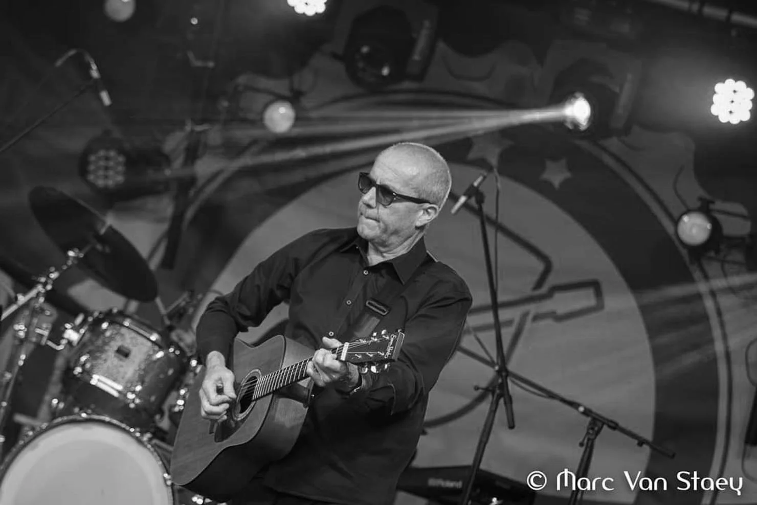 Man speelt gitaar op een podium, met een drumstel op de achtergrond, in zwart-wit.